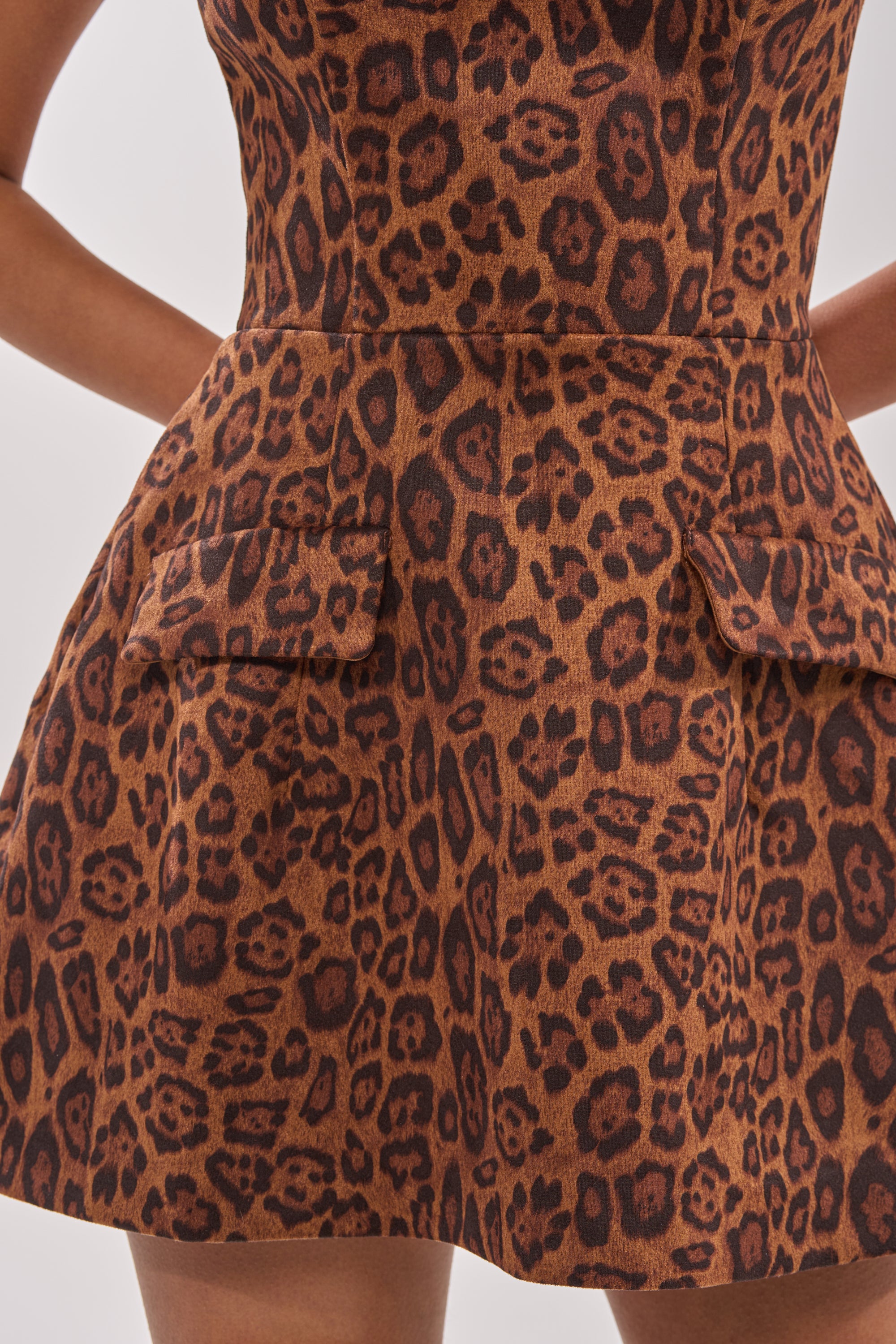 A close-up of a person wearing the CHEETAH GIRL MINI DRESS—a brown mini dress with bold cheetah print, dark spots, and two front flap pockets—standing with hands behind their back. 6