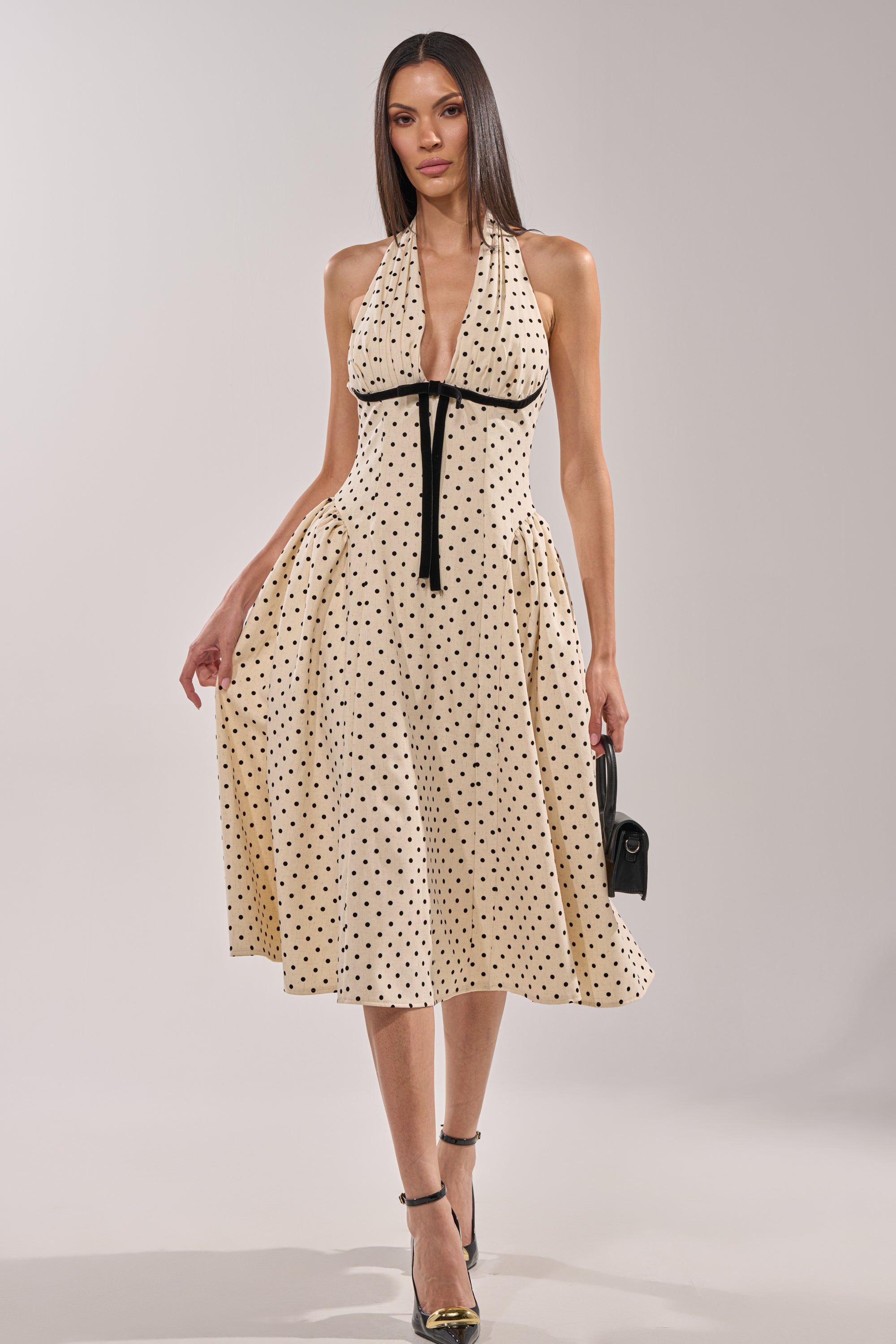 A woman wears the SUNDAY AFFAIR MAXI DRESS, a cream halter-neck maxi with black polka dots and a black tie under the bust, styled with black heels and a small black handbag. The vintage-inspired look is set against a plain, light background. 0