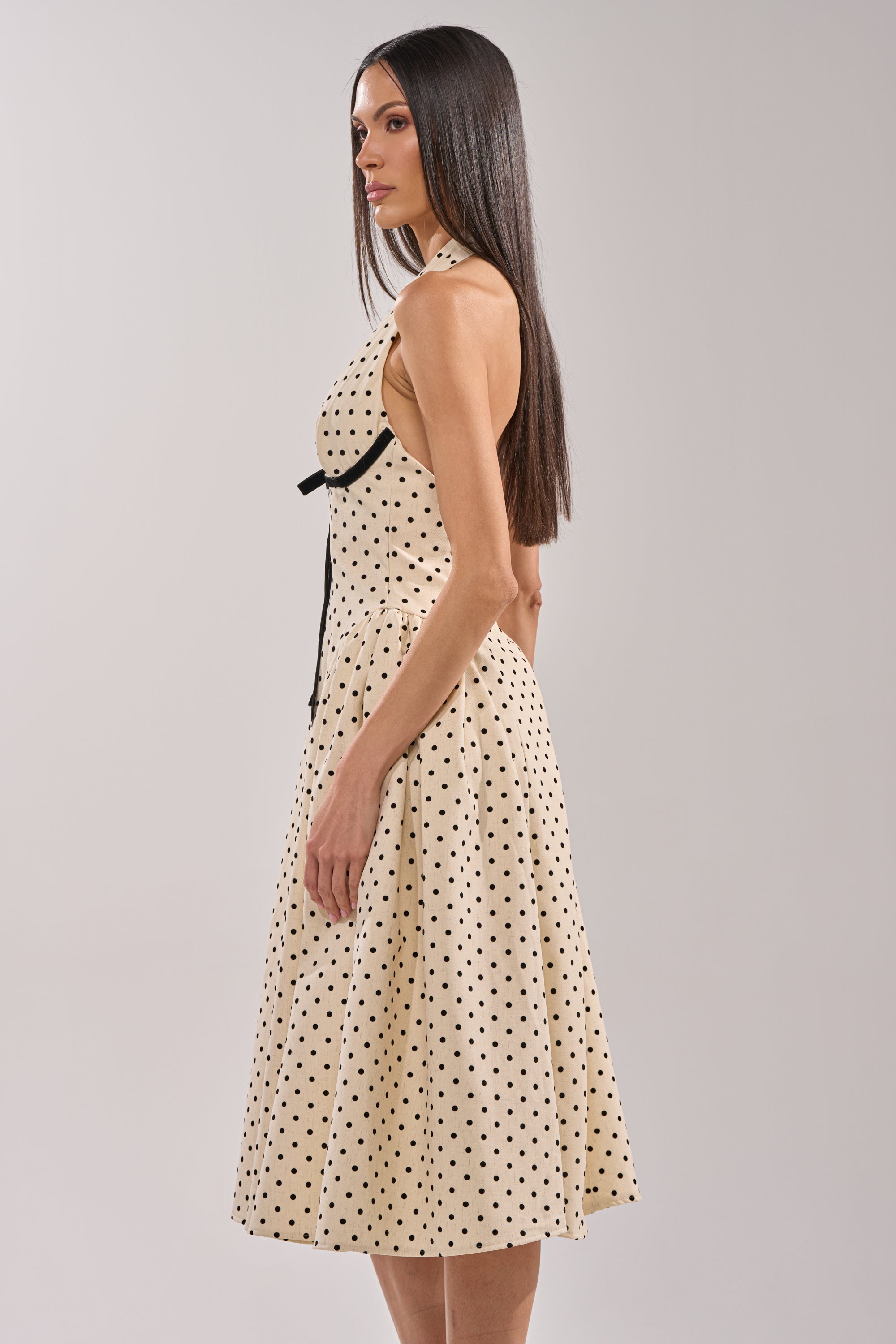 A woman with long straight hair stands in profile against a plain light background, wearing the SUNDAY AFFAIR MAXI DRESS—a vintage-inspired cream polka dot halter dress with black ribbon detail. 3