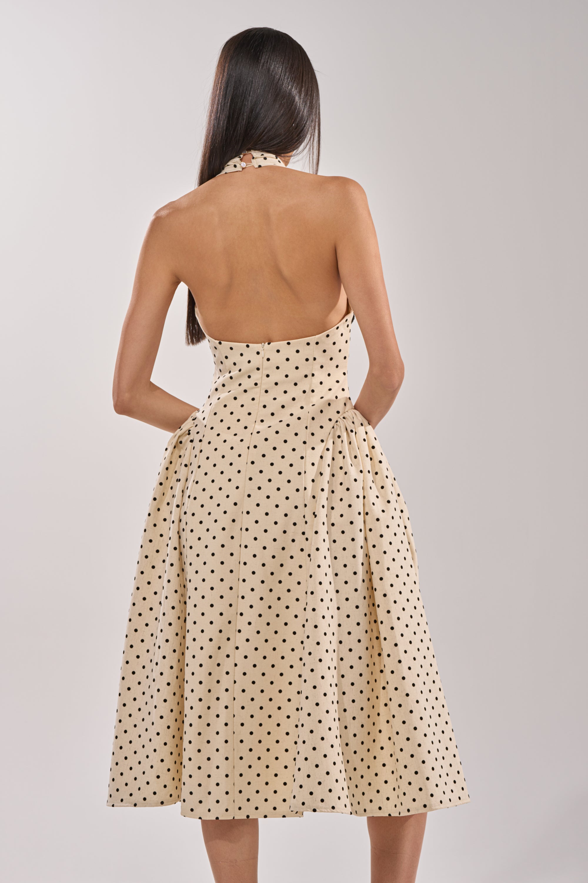 A woman with long dark hair wears the SUNDAY AFFAIR MAXI DRESS—a vintage-inspired beige halter neck maxi dress with black polka dots—posing with her back to the camera against a plain light background. 4
