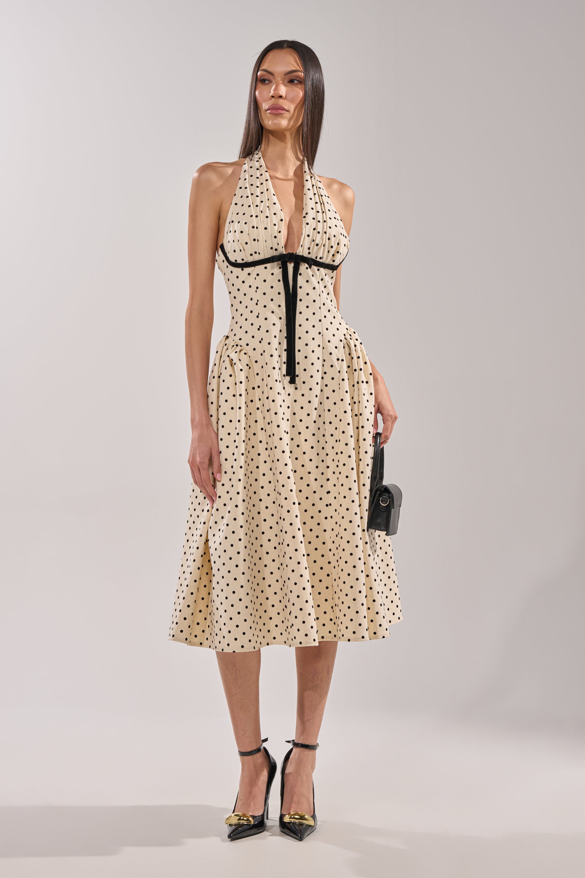 A woman wears the SUNDAY AFFAIR MAXI DRESS featuring a vintage-inspired halter neck, black polka dots, and a black ribbon under the bust. She pairs it with black heels and a small black handbag, standing against a plain light background. 5