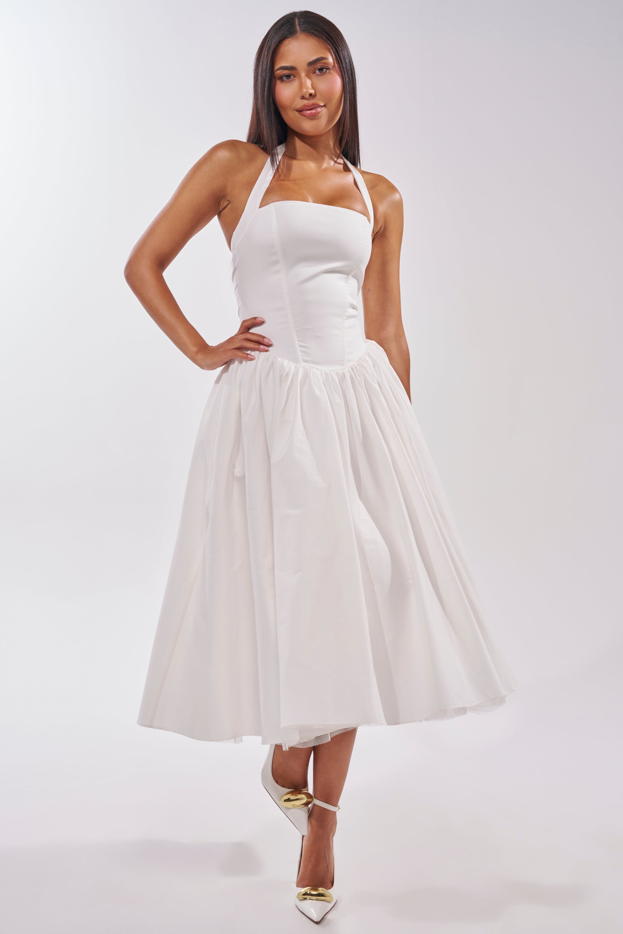 A woman with straight dark hair wears the SUNDAY AFTERNOON POPLIN MIDI DRESS IN WHITE, featuring a fitted bodice and flared skirt—ideal for garden parties or bridal events. She completes the look with metallic pointed-toe heels. 1