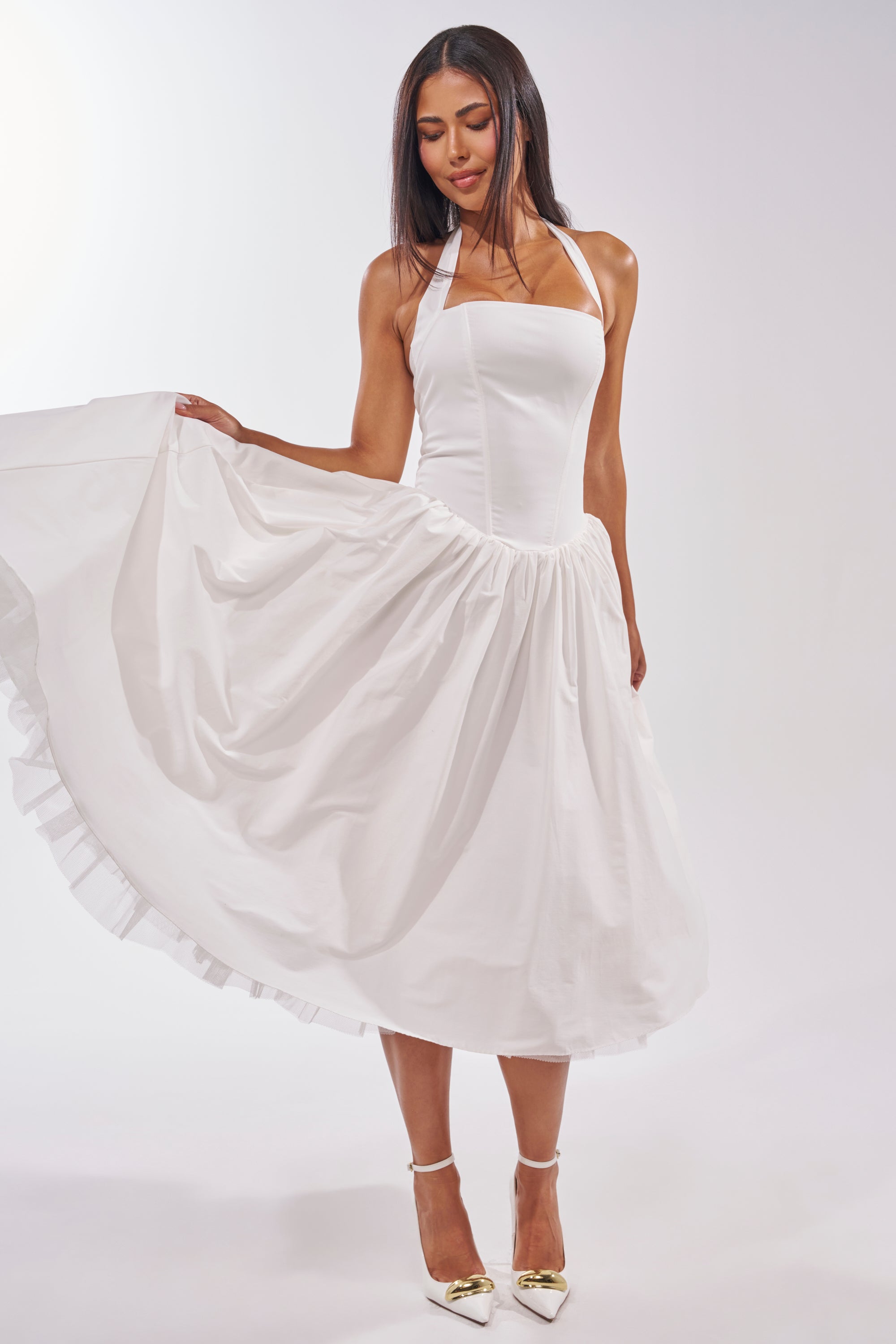 A woman in the SUNDAY AFTERNOON POPLIN MIDI DRESS IN WHITE holds out one side of her skirt, ideal for garden parties or bridal events. She has long dark hair, a slight smile, and wears white heels with gold tips against a plain light background. 2
