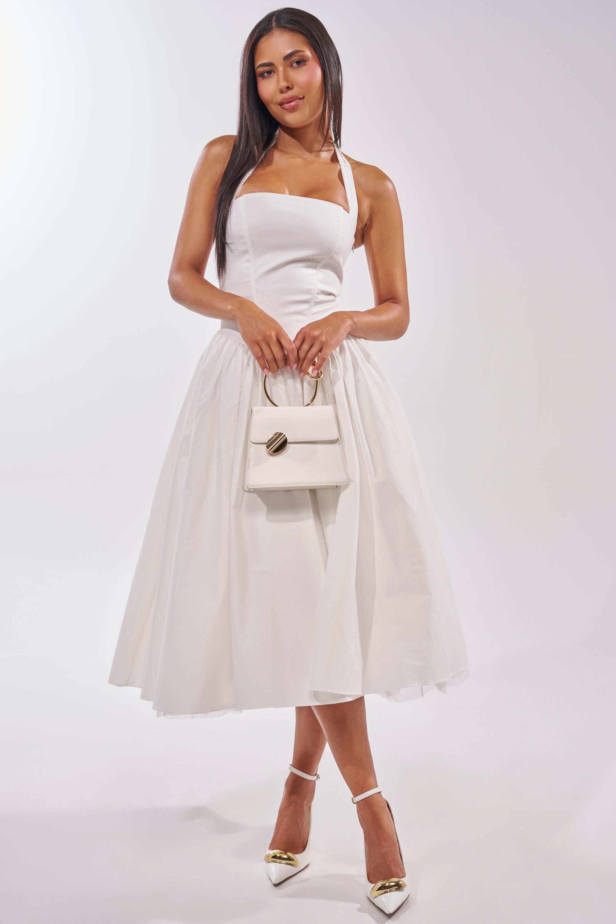 Wearing the SUNDAY AFTERNOON POPLIN MIDI DRESS IN WHITE, a woman holds a small white handbag and wears pointed white heels with gold accents. Perfect for bridal events or garden parties, she stands against a light background, looking at the camera. 3