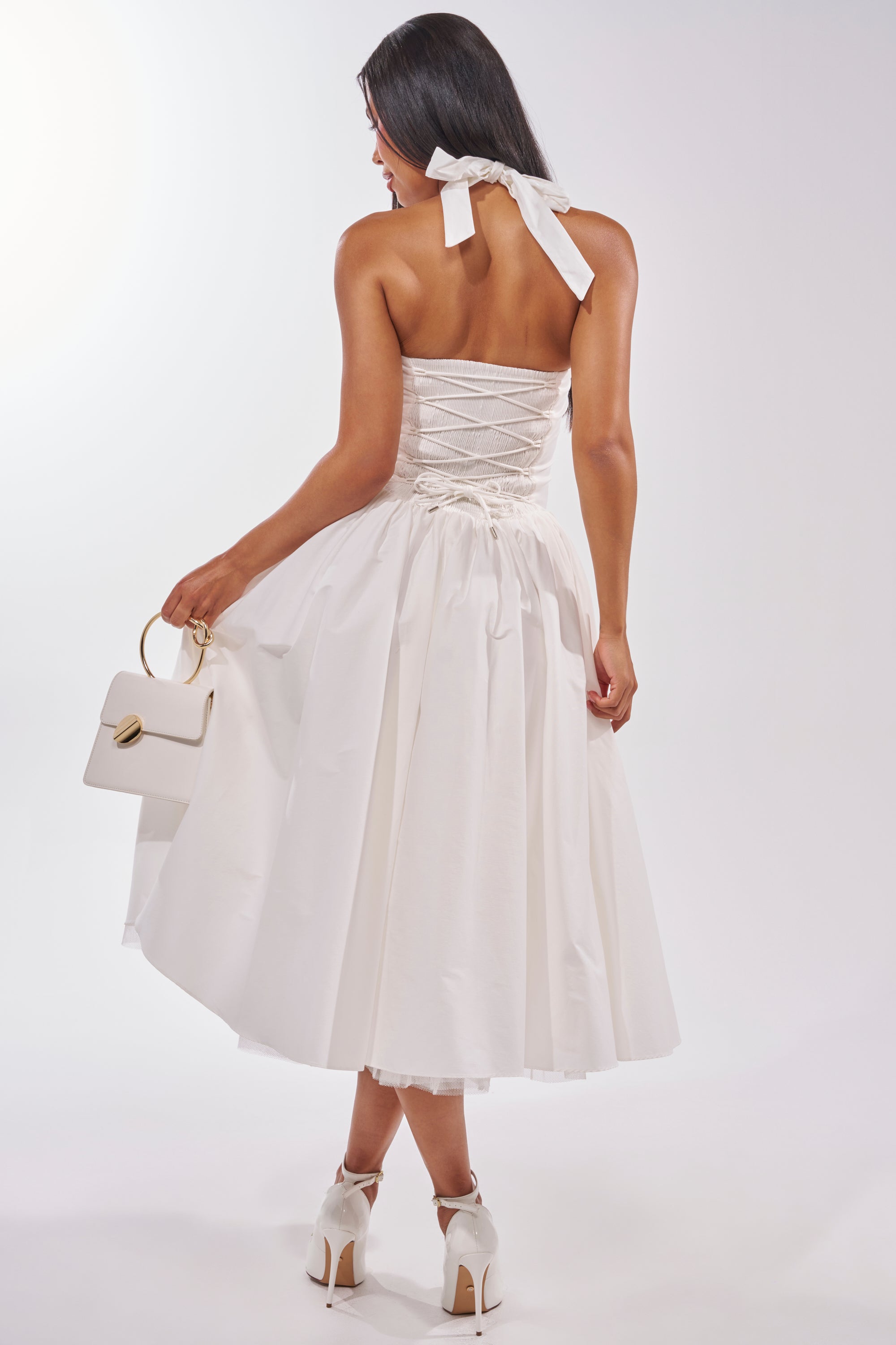 A woman in the SUNDAY AFTERNOON POPLIN MIDI DRESS IN WHITE stands facing away, holding a white handbag in her left hand. She pairs the dress with white heels and ties her long dark hair back with a ribbon. 5