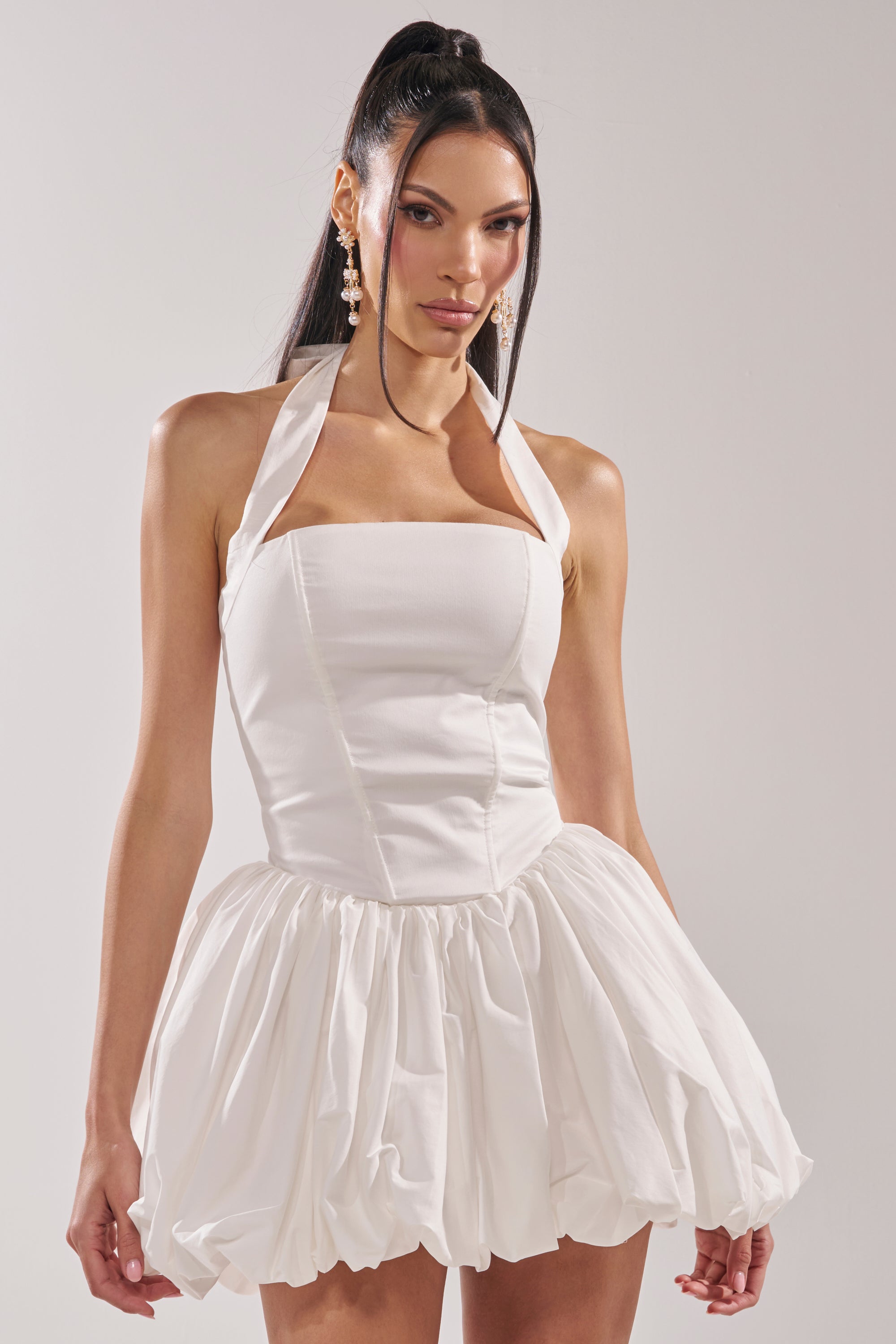 A woman in the BALLERINA POPLIN MINI DRESS IN WHITE—with a fitted corset bodice and bubble skirt—stands against a plain light background. Her long dark hair is in a high ponytail, and she completes the look with dangling earrings. 1