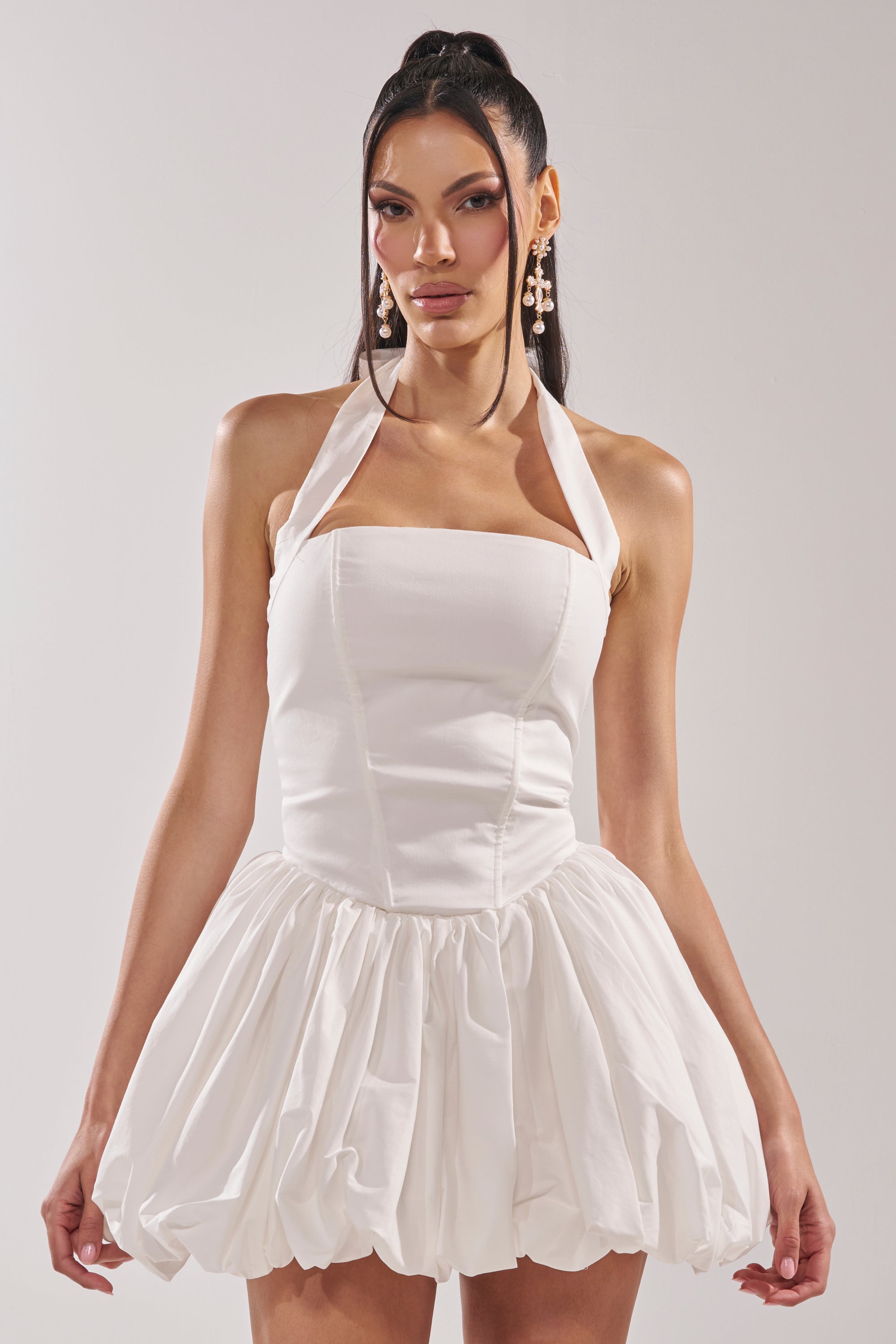 A woman models the BALLERINA POPLIN MINI DRESS IN WHITE, featuring a corset bodice and voluminous bubble skirt. She styles it with a high ponytail and statement earrings, posing against a plain background. 2