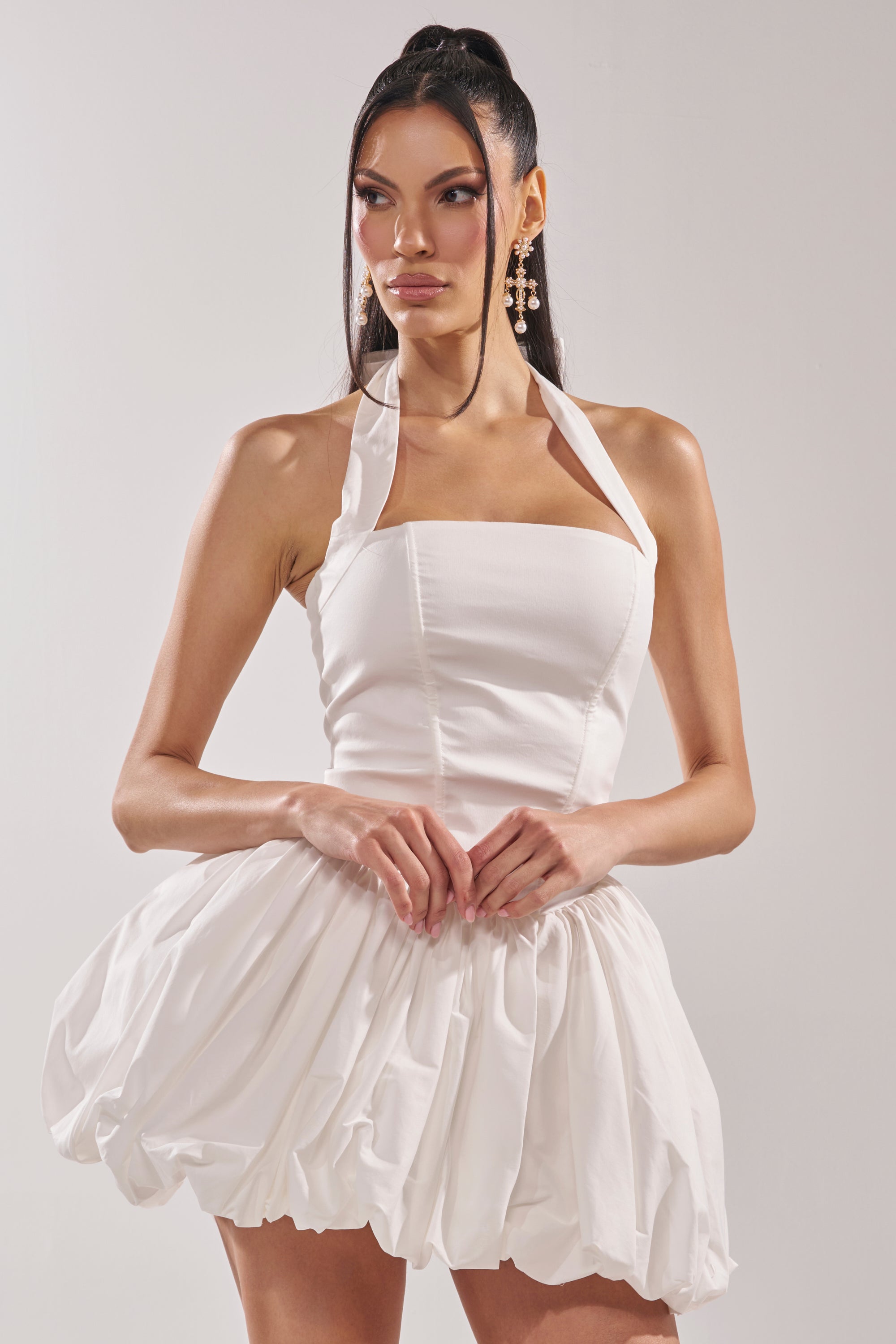 A woman with long dark hair in a high ponytail and statement earrings models the BALLERINA POPLIN MINI DRESS IN WHITE, featuring a corset-inspired bodice and voluminous bubble skirt, against a plain background. 3
