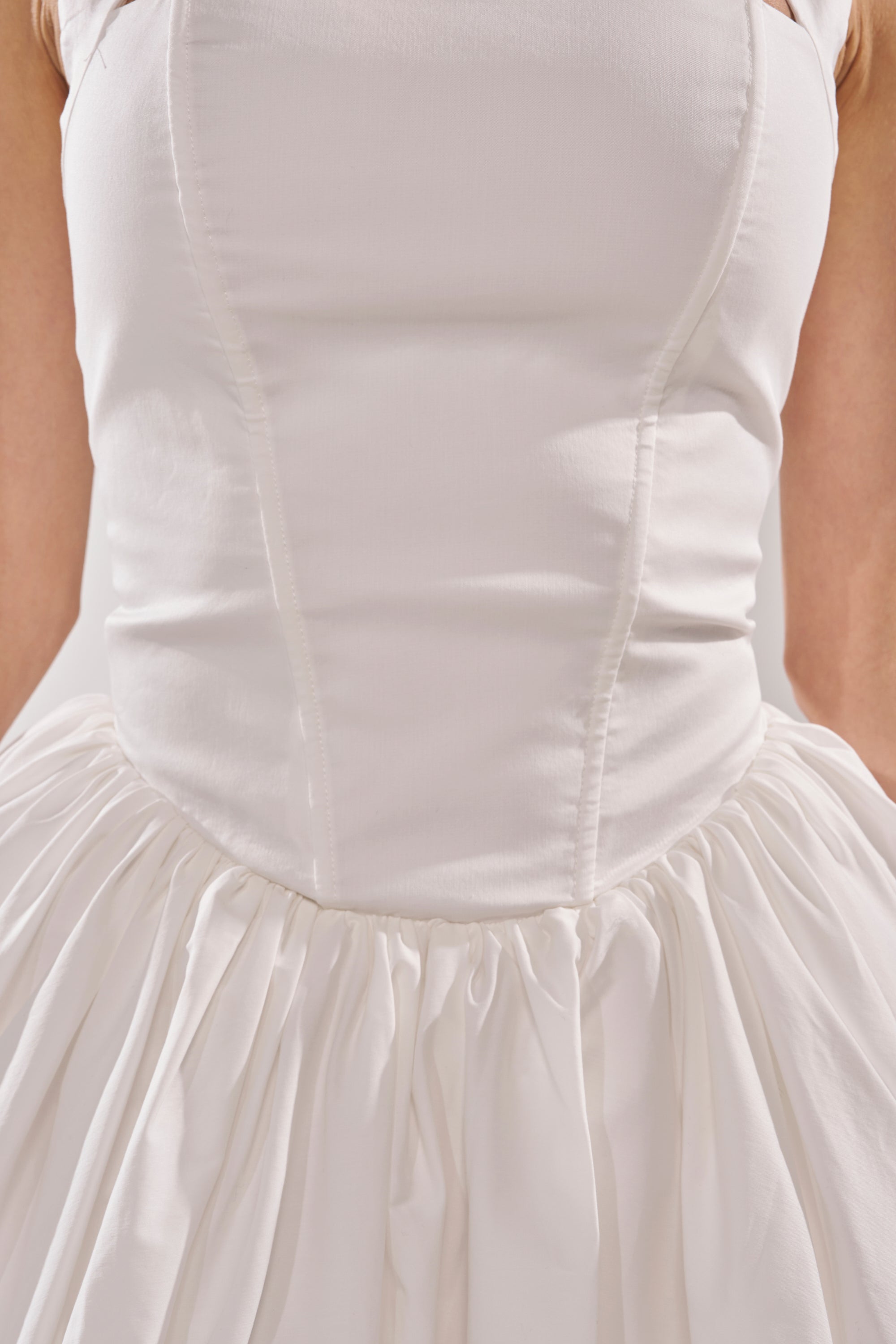 A close-up of a person wearing the BALLERINA POPLIN MINI DRESS IN WHITE, featuring a fitted bodice and gathered, flowing skirt. The image highlights the torso and part of the skirt, with arms partially visible. 8