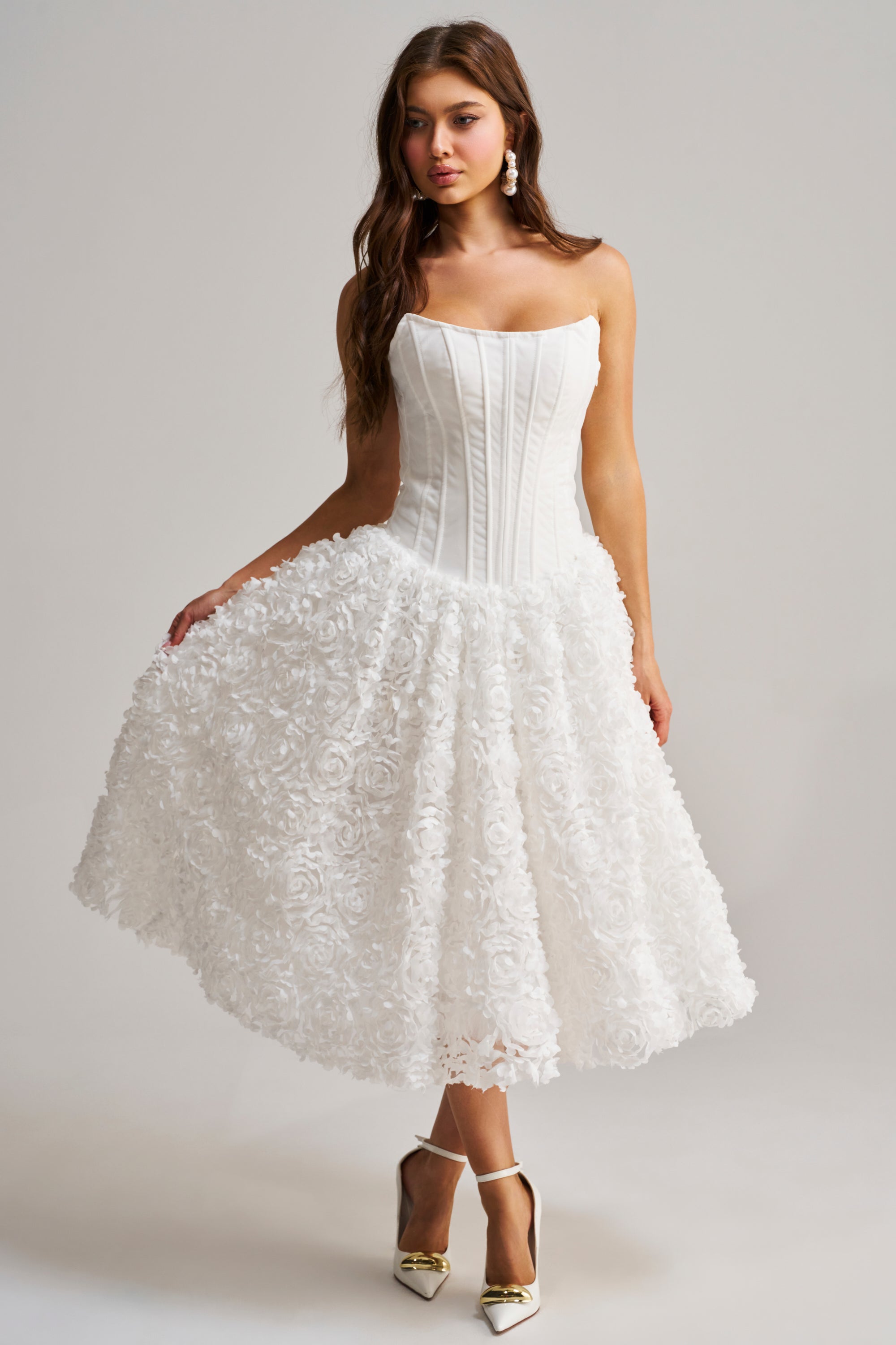 A woman wearing the FIND ME LATER FLORAL CORSET MIDI DRESS IN WHITE, featuring a fitted bodice and textured floral knee-length skirt, paired with gold heels and pearl earrings, stands against a plain light gray background.