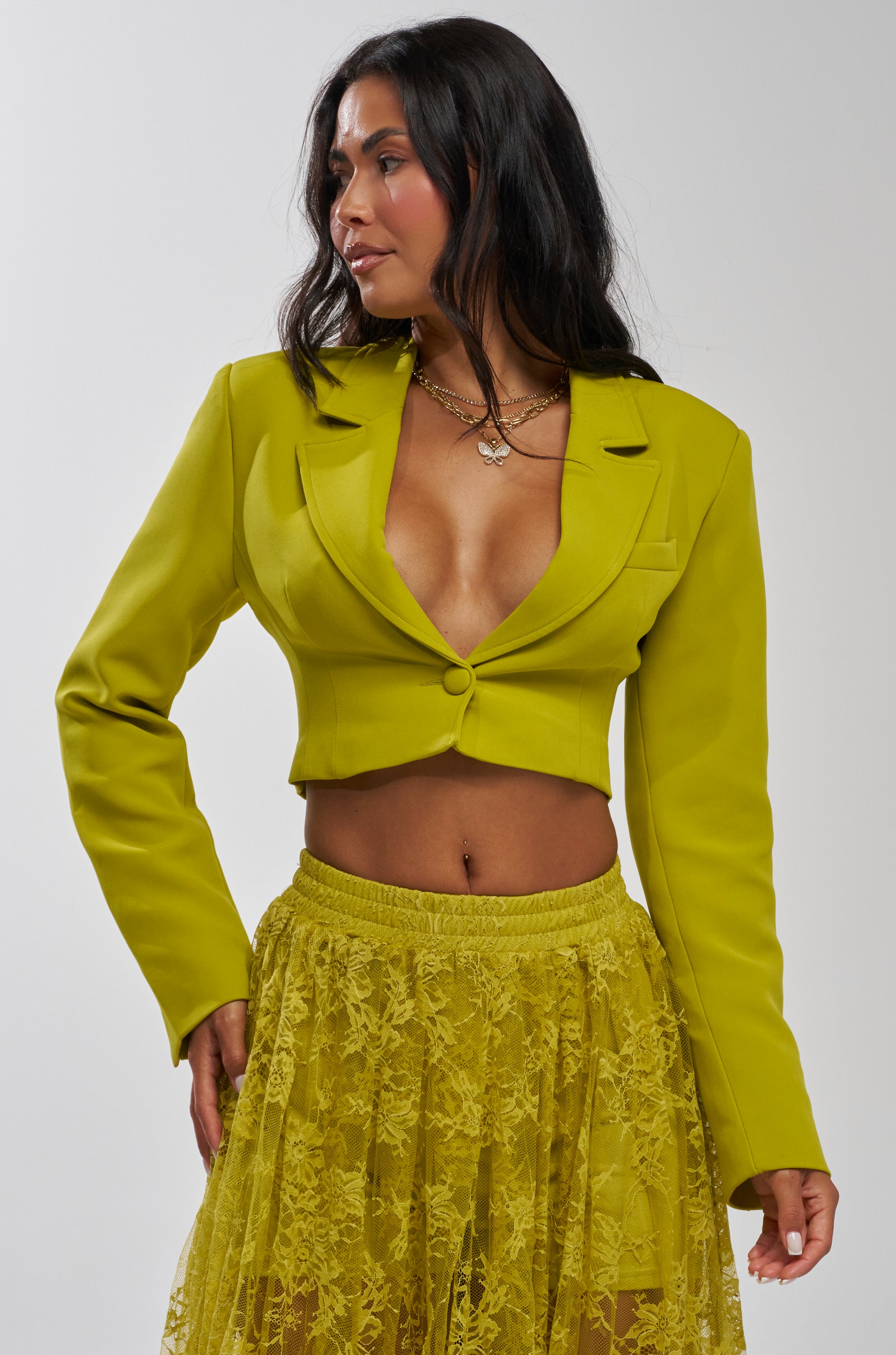 A woman poses against a plain light gray background wearing the CAN I KICK IT CROPPED BLAZER in chartreuse with a deep neckline, styled with a matching sheer lace skirt.