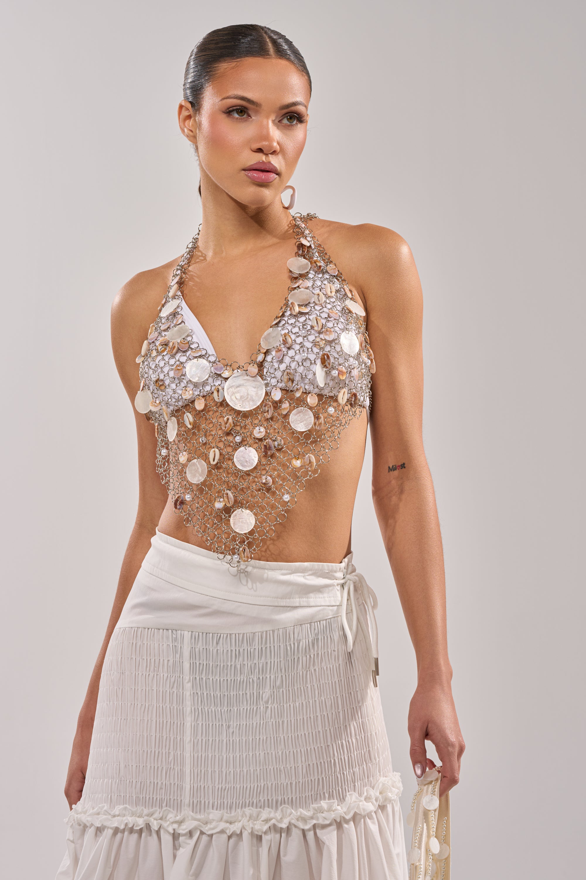A woman wears the SALLY SELLS SEASHELLS CROCHET HALTER TOP over a white bandeau and a long, pleated white skirt, standing against a light background—an elegant look for evening festival wear. 0