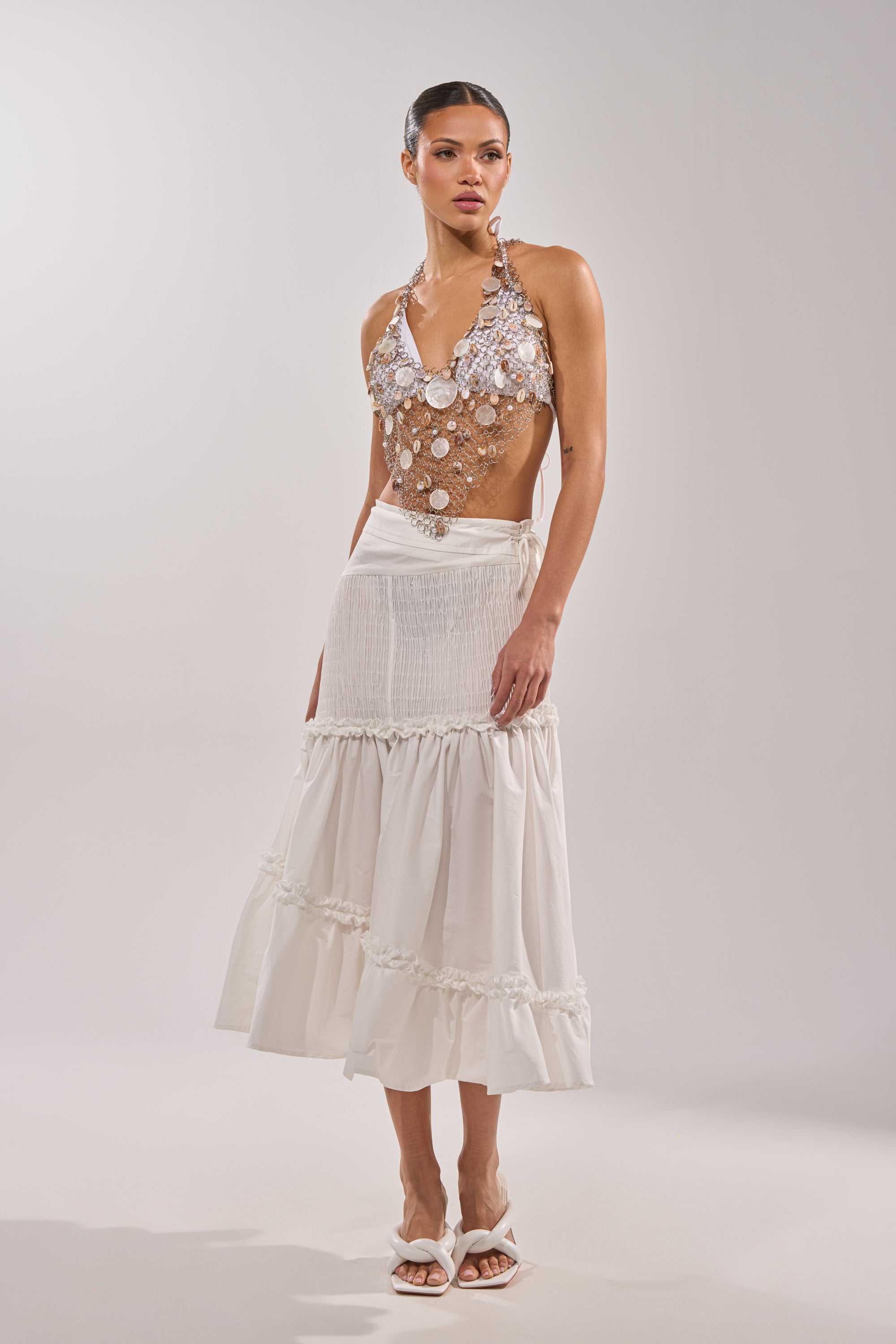 A woman wears the SALLY SELLS SEASHELLS CROCHET HALTER TOP with metallic and shell-like discs, paired with a white ankle-length skirt and sandals—an eye-catching festival look against a plain light background. 5