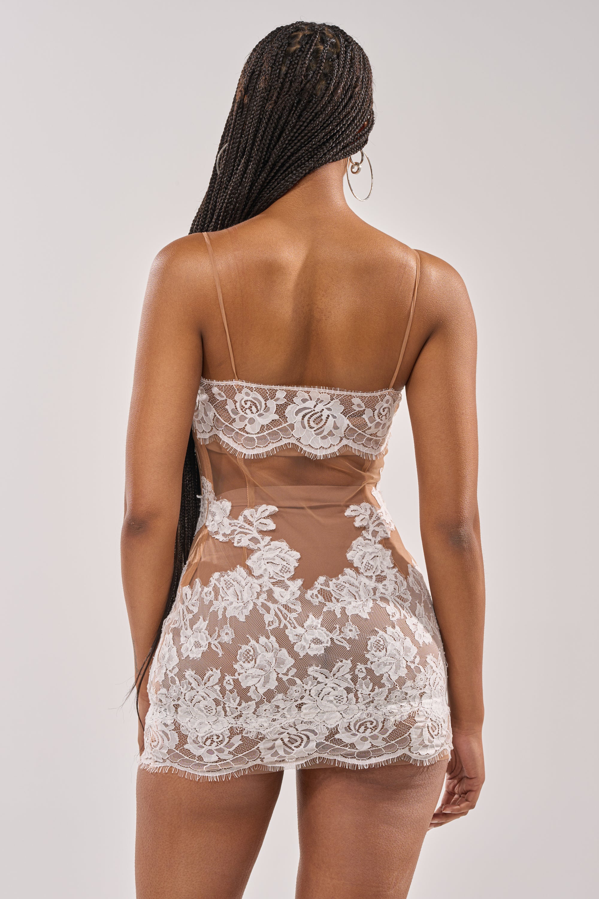 A woman with long braided hair faces away, modeling the LOVING ON YOU LACE MINI DRESS in nude and ivory. This sheer lace mini dress features white floral patterns, thin straps, a fitted silhouette, and delicate detailing. 6