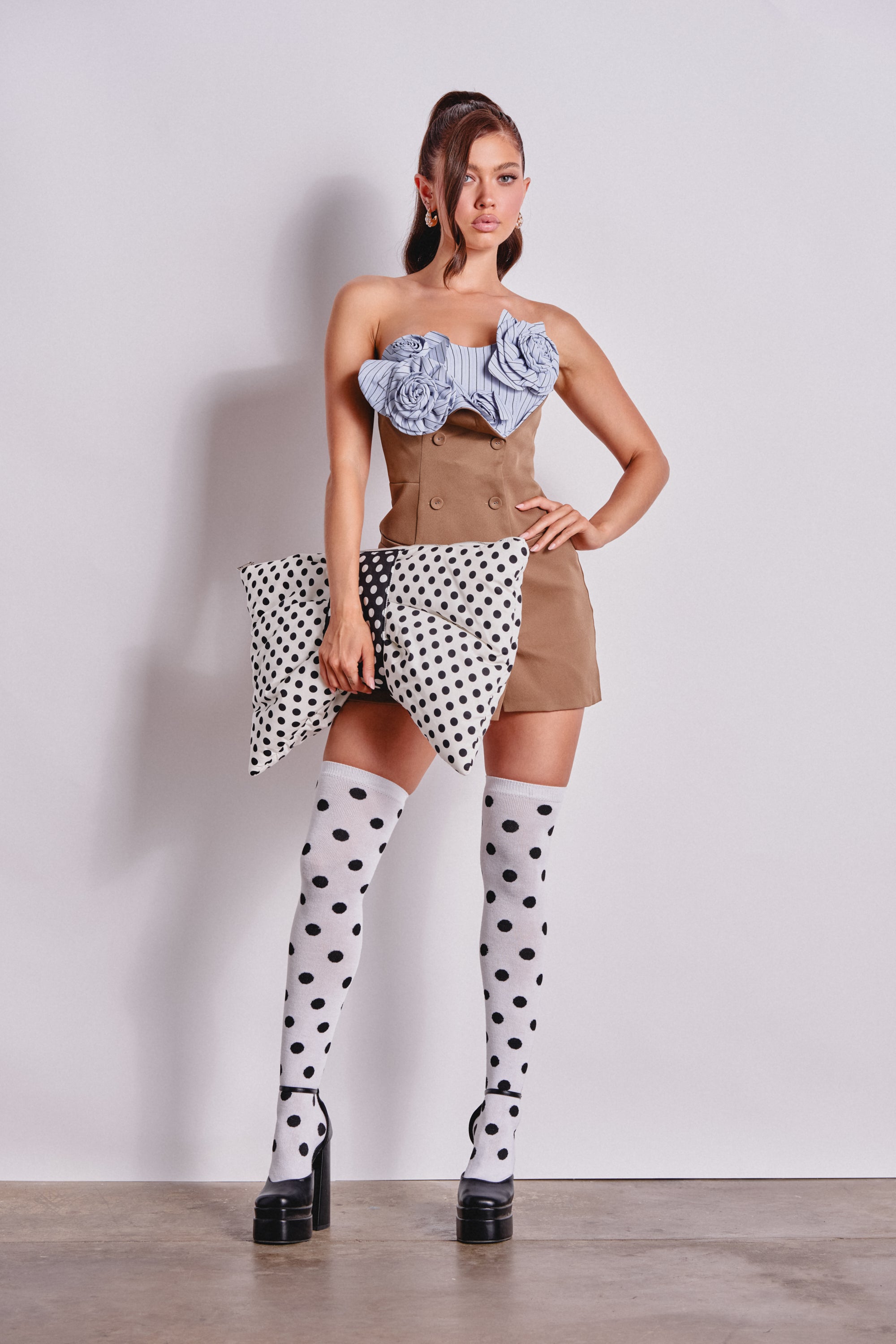 A woman poses against a plain wall, wearing the OH DARLING FLORAL APPLIQUE POPLIN MINI DRESS, paired with polka dot thigh-high socks, platform heels, and holding a large polka dot clutch. 6