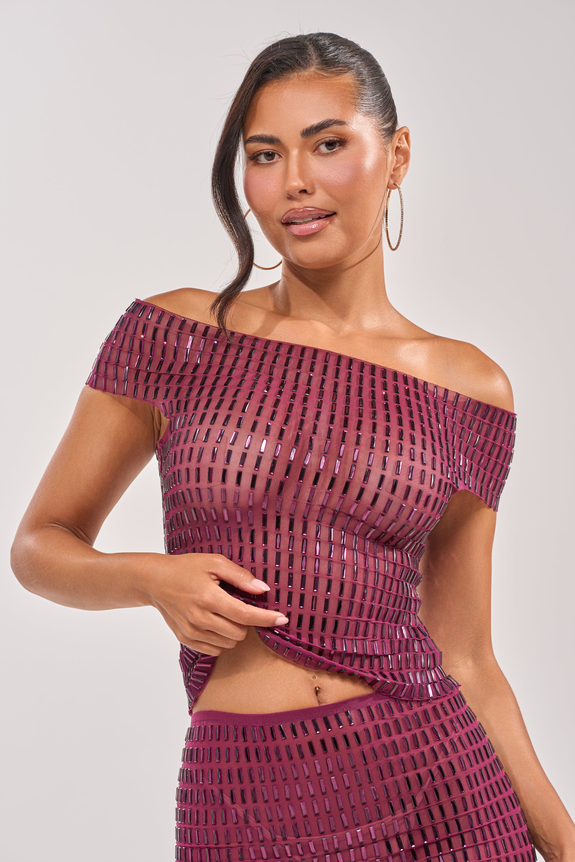 A woman poses in the SHAKE IT FOR ME BABY RHINESTONE TOP, an off-the-shoulder, shiny party top with a geometric pattern. She has long dark hair to the side, wears large hoop earrings, and stands before a plain light background. 4