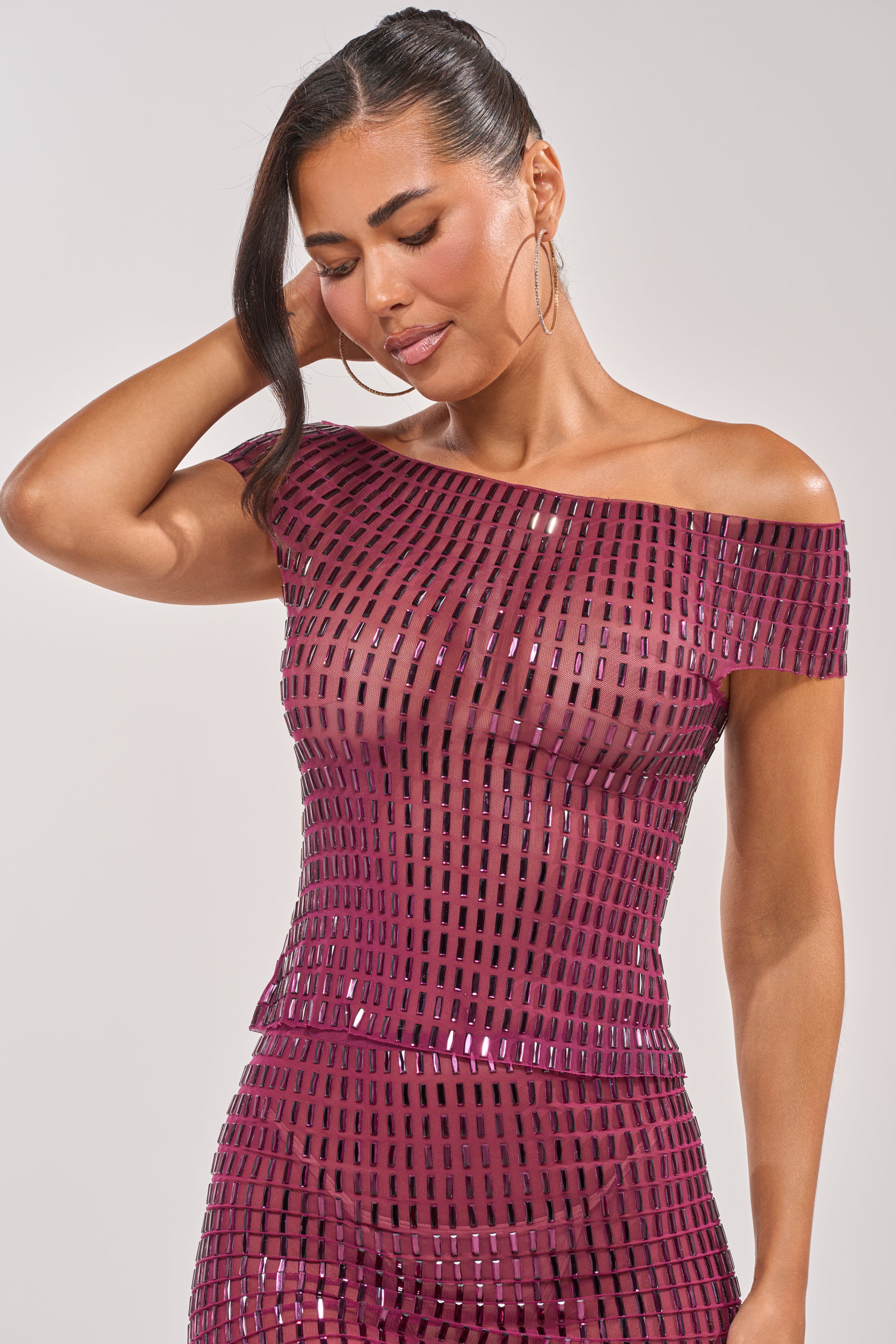 A woman smiles softly with eyes closed in the SHAKE IT FOR ME BABY RHINESTONE TOP, a shiny, off-the-shoulder party piece with a grid pattern, posing with one hand in her hair against a plain background. 5