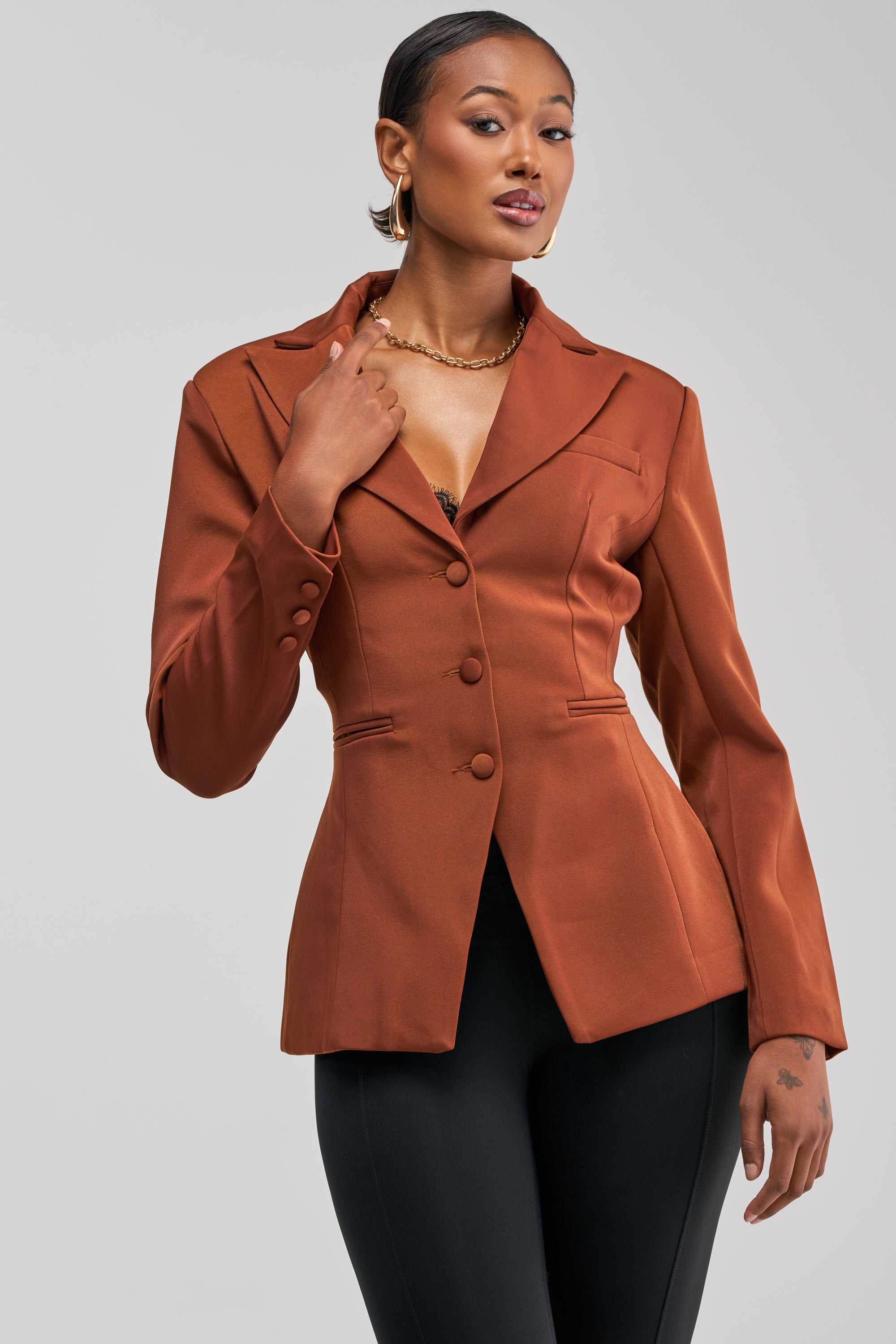 A woman poses confidently against a plain light background, showcasing effortless women’s workwear style in the rust-colored CATALINA BLAZER with buttons and black pants.