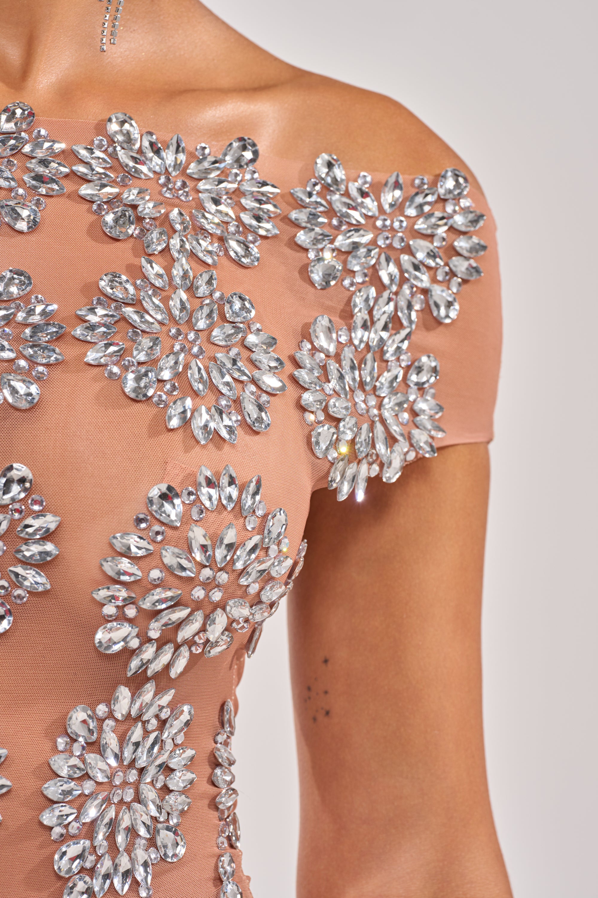 A close-up of a person wearing the SO TEMPTING RHINESTONE OFF SHOULDER MINI DRESS in peach, featuring large silver rhinestone floral patterns on mesh, reveals some skin and a small upper arm tattoo. 9