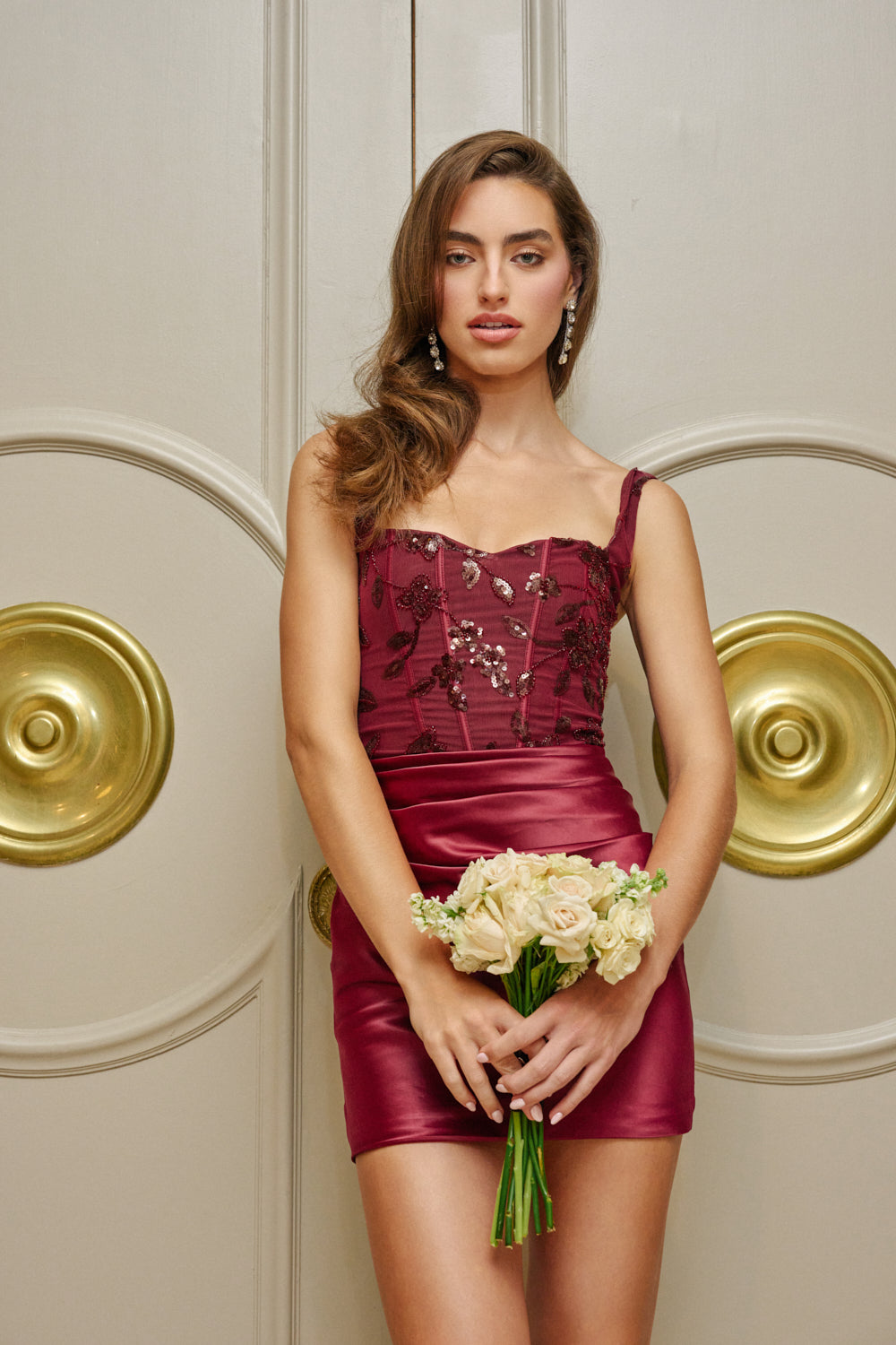 Wearing the HEARTBREAKER CORSET MINI DRESS, a woman in a burgundy mesh dress adorned with floral details holds a bouquet of pale roses as she stands before ornate double doors with gold circular accents—the perfect choice for an elegant homecoming outfit.
