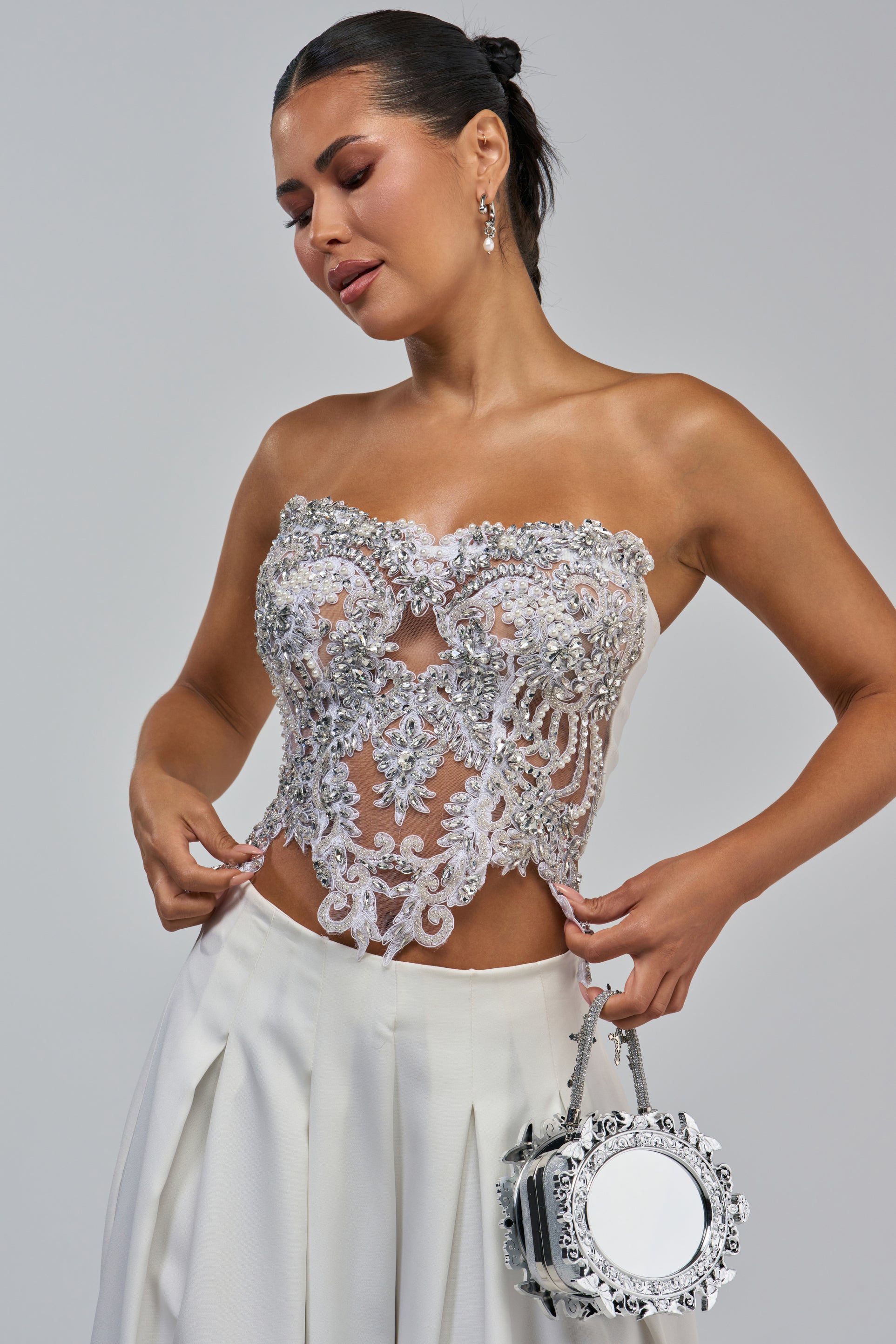 Dressed in the LET’S GET MARRIED EMBELLISHED CORSET paired with white pleated pants, a woman holds a round silver purse as she poses against a plain, light background, looking down with a gentle smile.