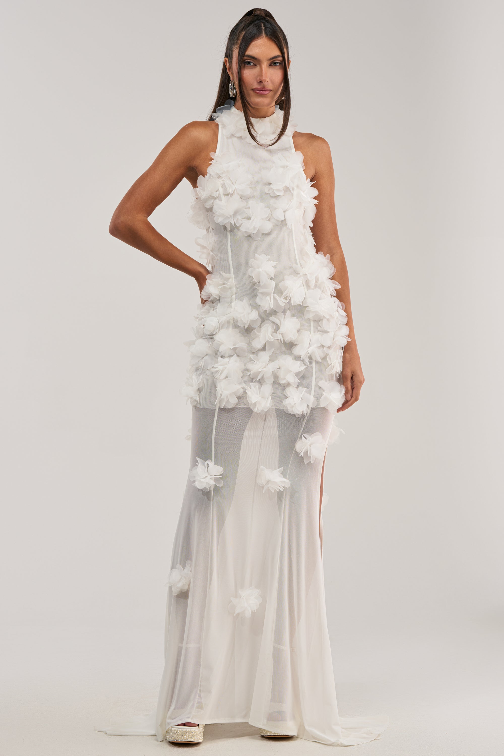A woman wears the PARTY FAVOR RUFFLE MAXI DRESS IN WHITE, a sleeveless formal maxi dress featuring 3D floral appliqués on the upper layer and a sheer skirt. She stands facing forward against a plain light background.