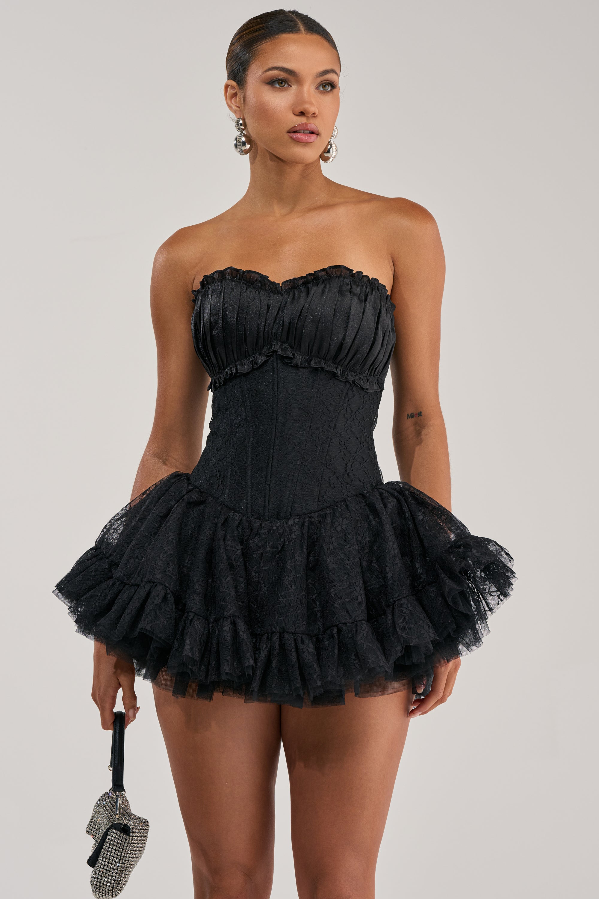 A woman models the PARTY STARTER CORSET LACE UP MINI DRESS, a strapless black dress with a ruffled tutu skirt. She accessorizes with a small silver handbag and dangling earrings, her hair pulled back against a light plain background.