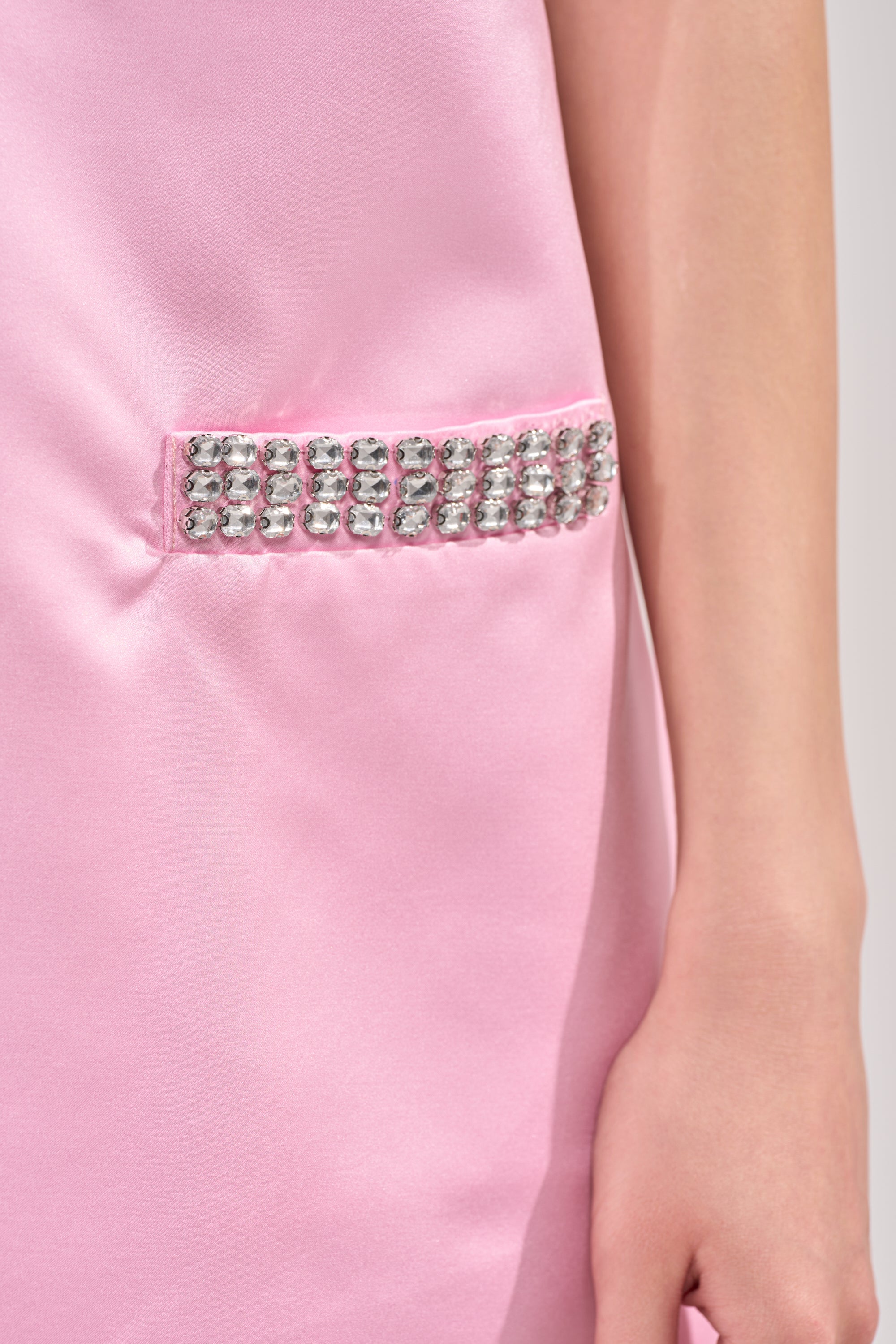A close-up of the UNDER THE LIGHTS SEQUIN MINI DRESS in pink satin, featuring a rectangular silver rhinestone embellishment near the waist. Only the wearer's arm and side are visible. 6