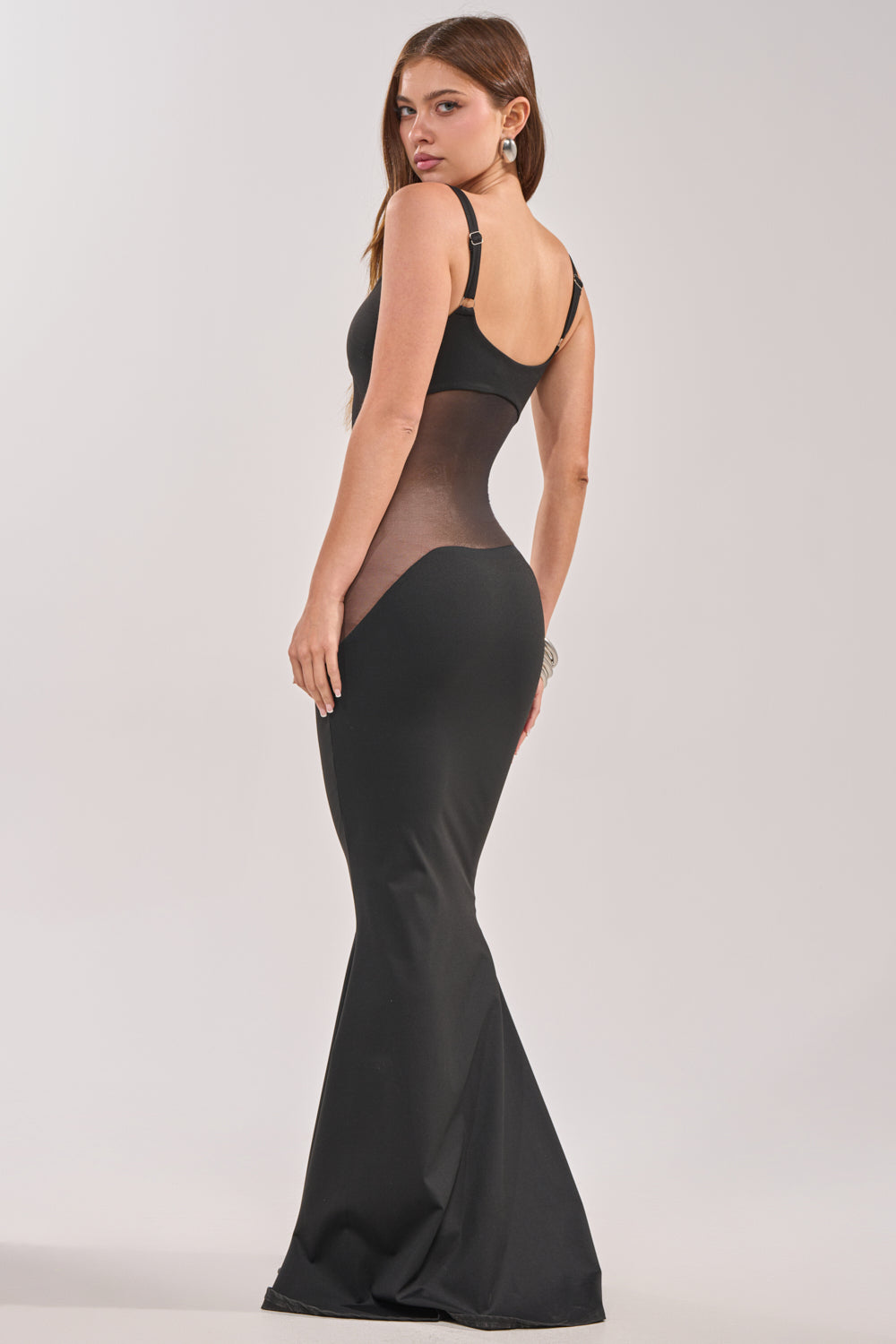 A woman poses in the SAY IT LOUDER MESH CUTOUT MAXI DRESS, a fitted floor-length style with sheer mesh side panels, standing with her back turned and looking over her shoulder against a plain background.