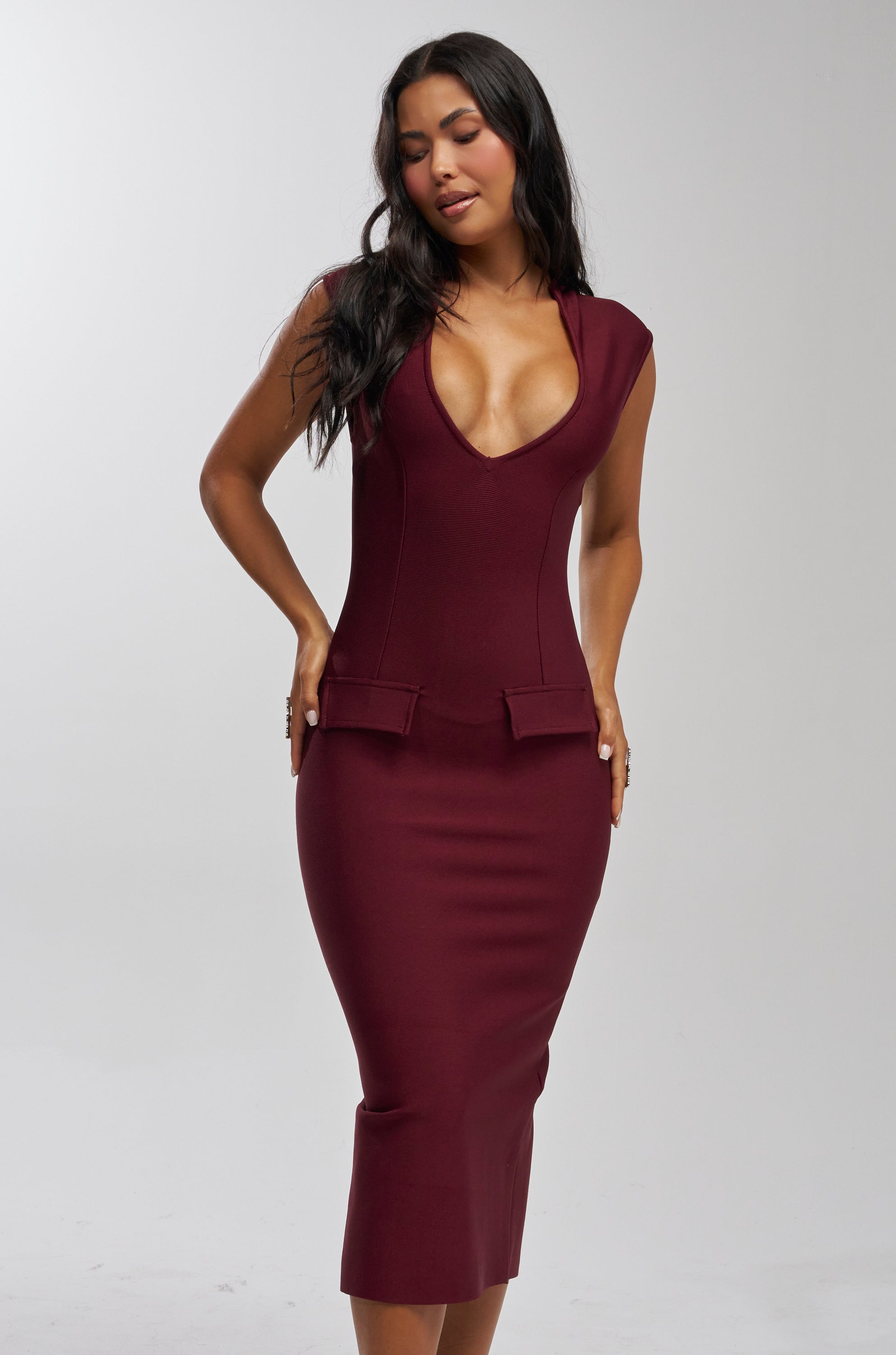 A woman with long dark hair models the KISS & TELL BANDAGE MIDI DRESS—a fitted, sleeveless burgundy midi dress with a deep V neckline and decorative pocket flaps—against a plain light background.