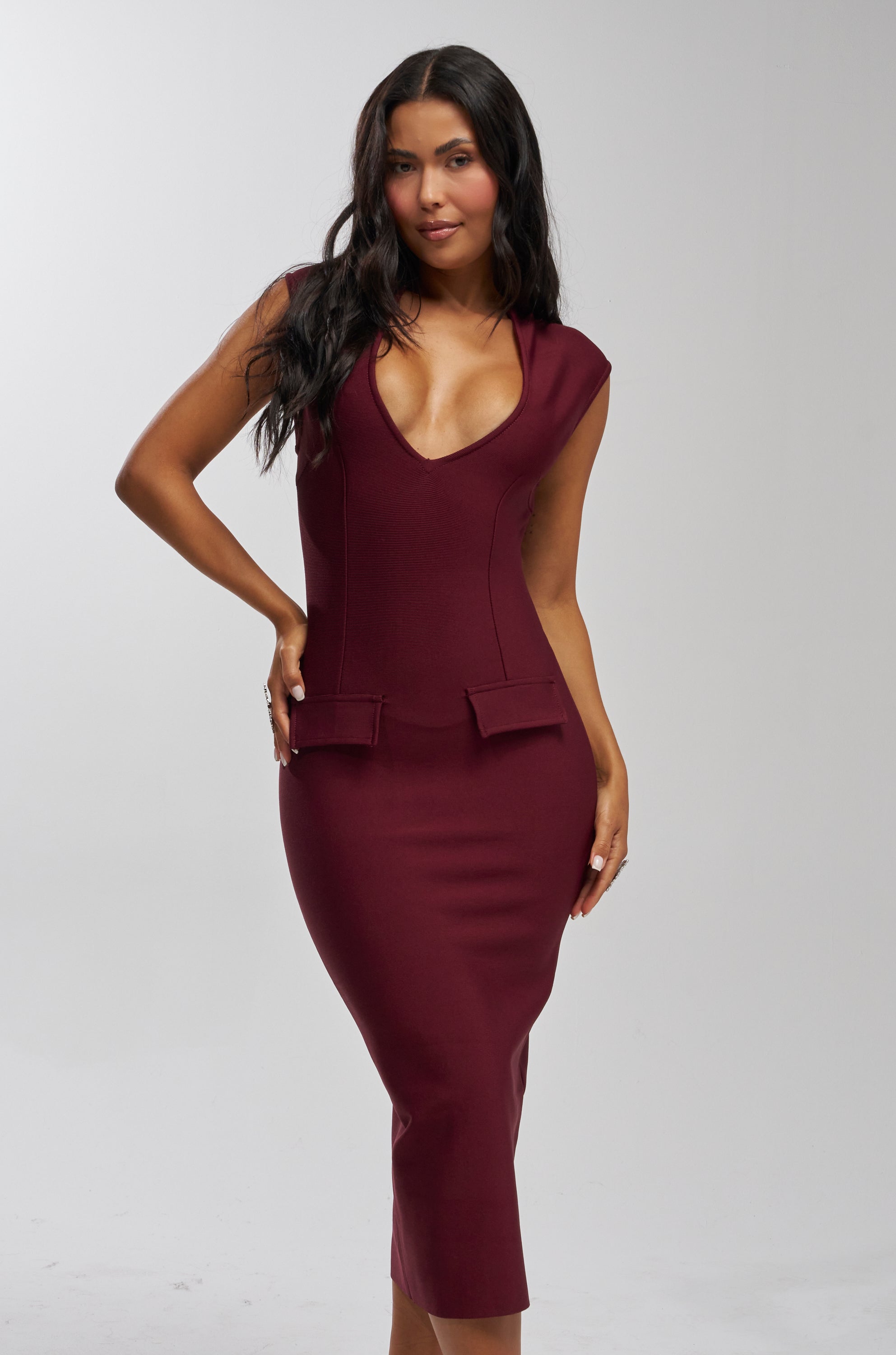 Woman modeling the KISS & TELL BANDAGE MIDI DRESS—a fitted, sleeveless burgundy midi dress with a deep V-neckline and decorative front pockets—stands against a plain background, making it a chic choice for your next cocktail party.