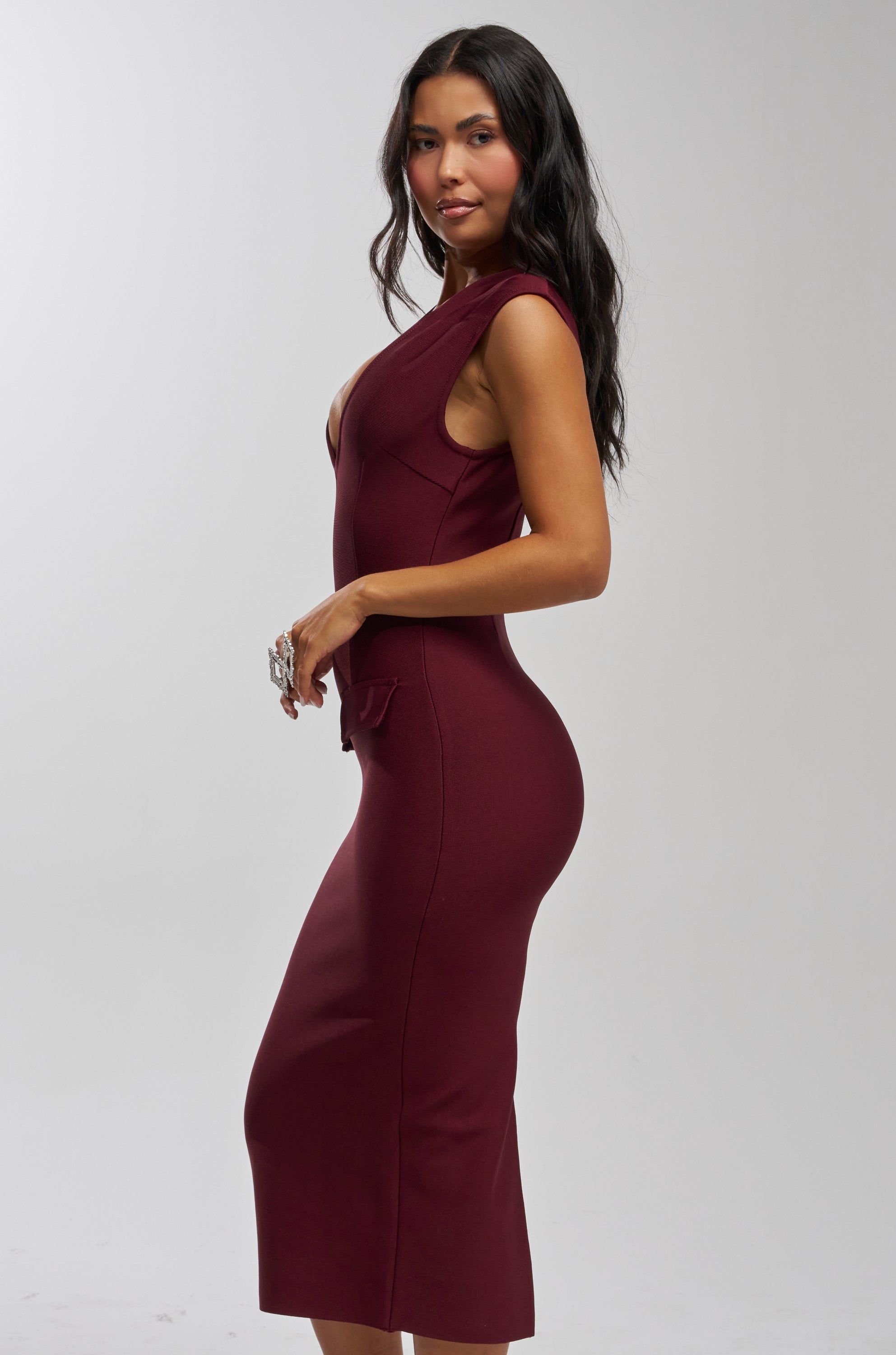A woman with long dark hair wears the KISS & TELL BANDAGE MIDI DRESS, standing sideways and smiling at the camera against a plain light background.