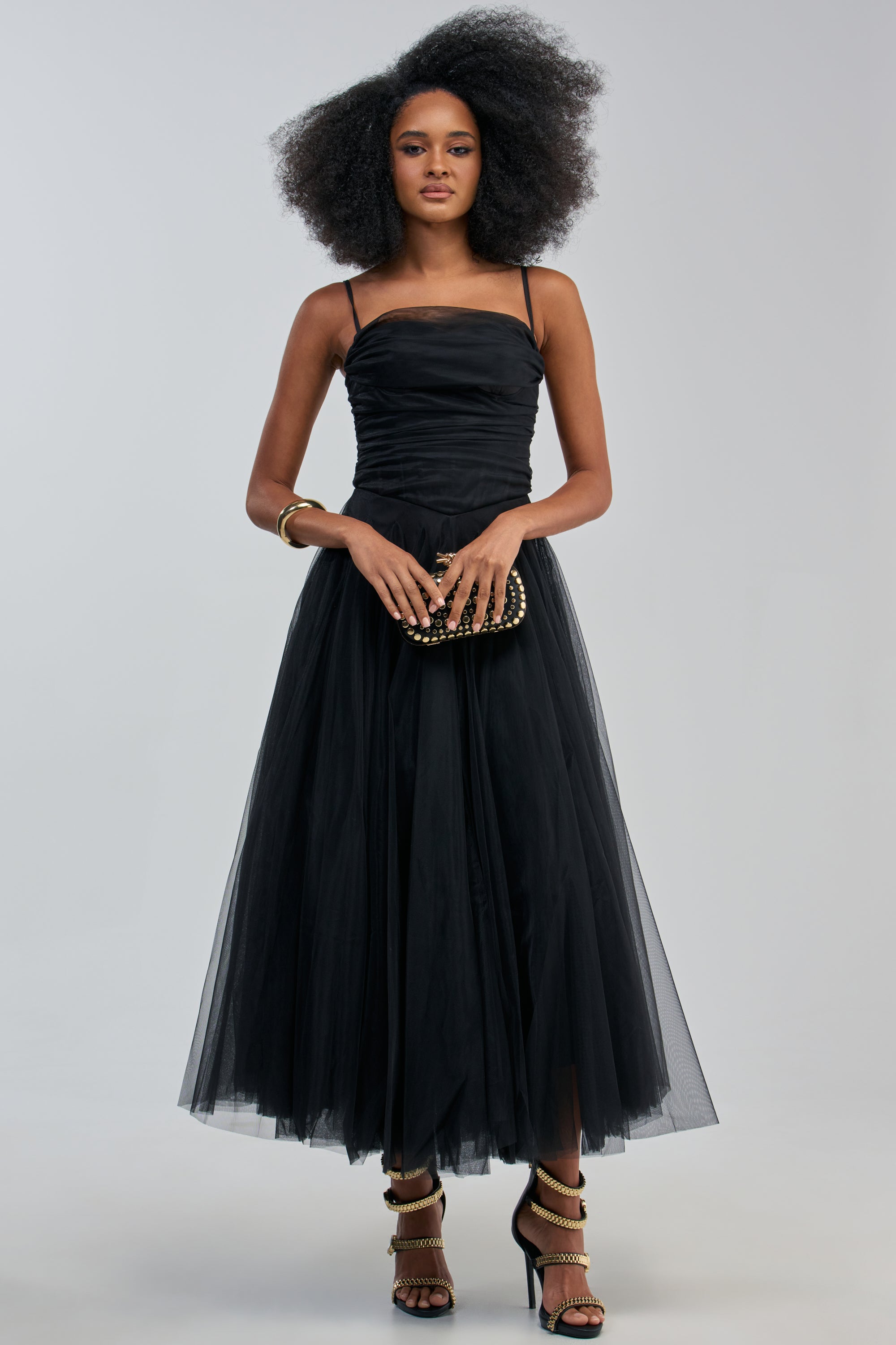 A woman in the SIMPLY STUNNING CORSET MAXI DRESS and strappy heels stands holding a black studded clutch. She accessorizes with a gold bangle and has voluminous, natural hair. The background is plain and light gray.