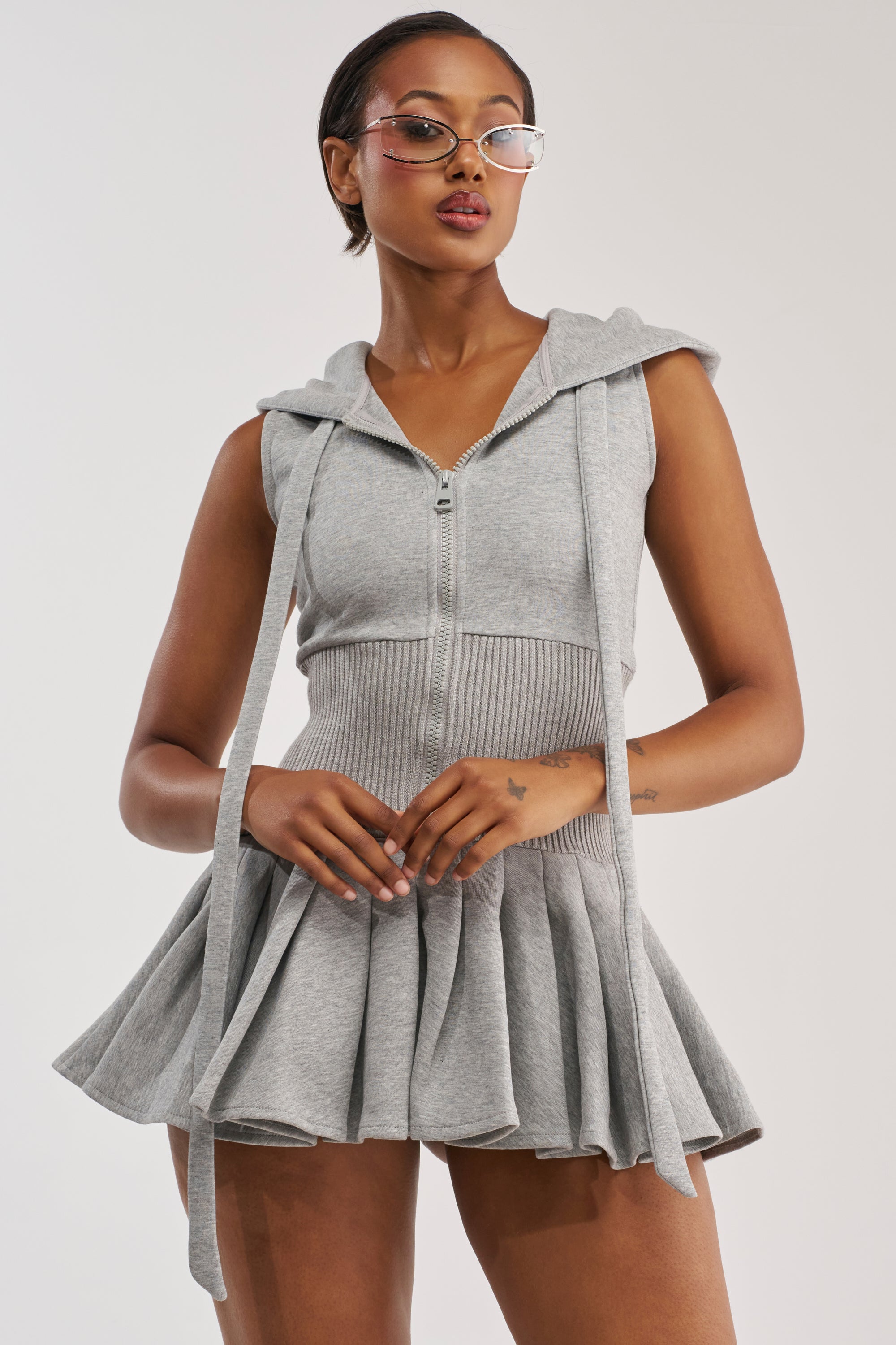 A woman wearing glasses poses against a plain white background in the REBEL WITH A CAUSE HOODED MINI DRESS, featuring a sleeveless, light gray zip-up design with a flared pleated mini skirt and long drawstrings for a sporty-chic look.