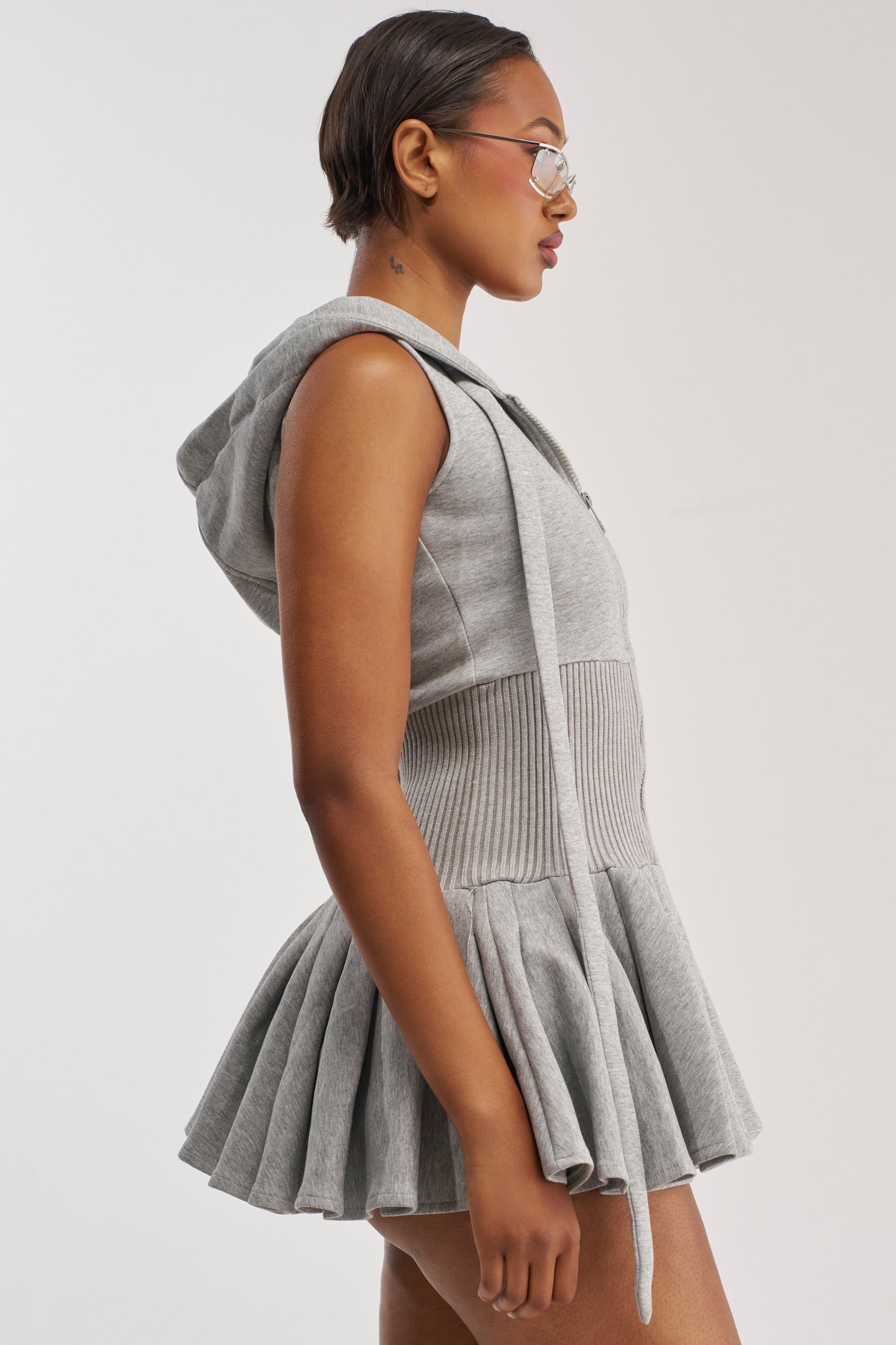 A person with short hair and glasses is seen in profile against a plain light background, wearing the REBEL WITH A CAUSE HOODED MINI DRESS—a sporty-chic, sleeveless grey mini dress featuring a pleated skirt and ribbed waist.