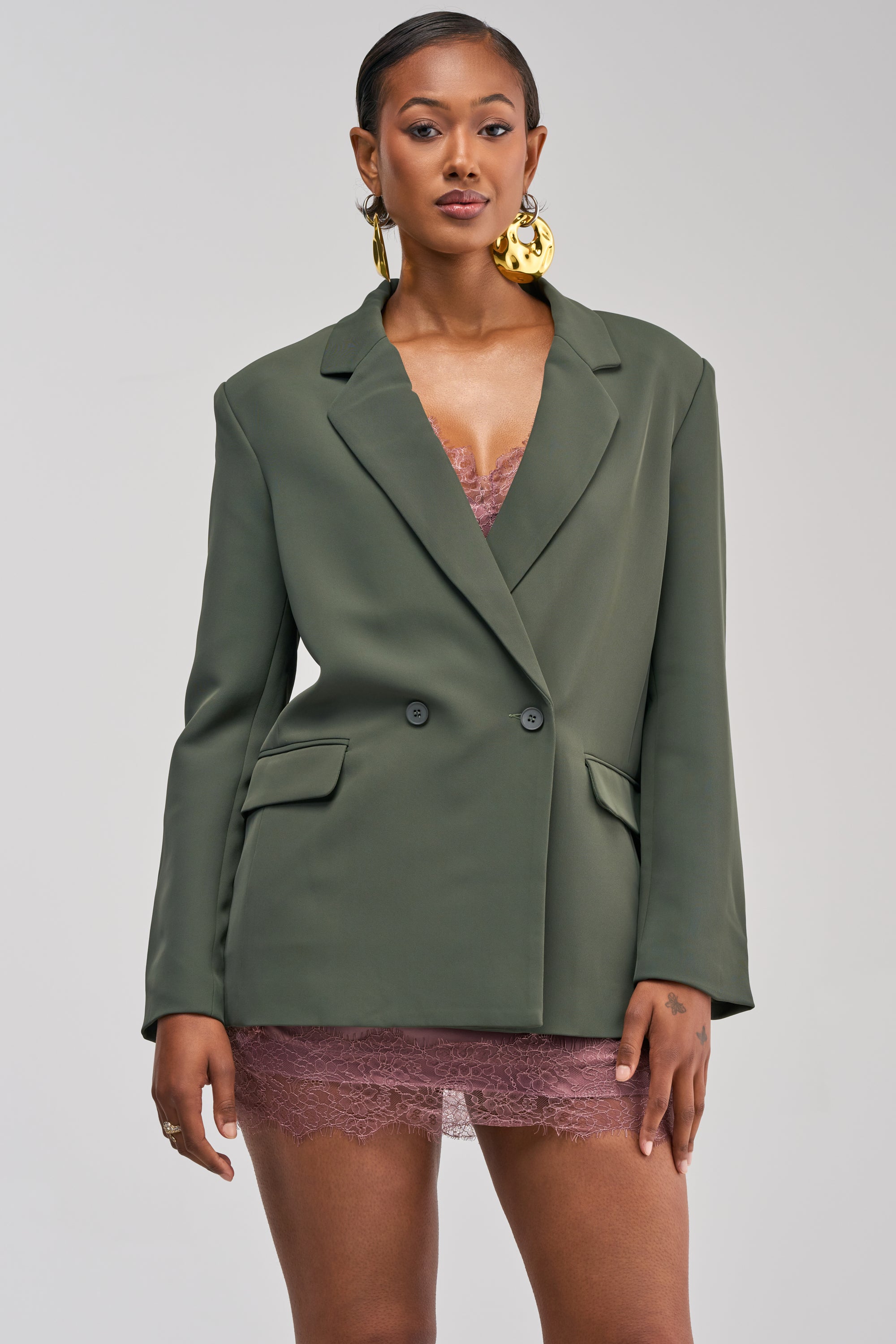 A woman stands against a plain light gray background, wearing the LOVE ME NOT BOYFRIEND BLAZER in oversized olive green layered over a mauve lace dress, and accessorized with large gold earrings.