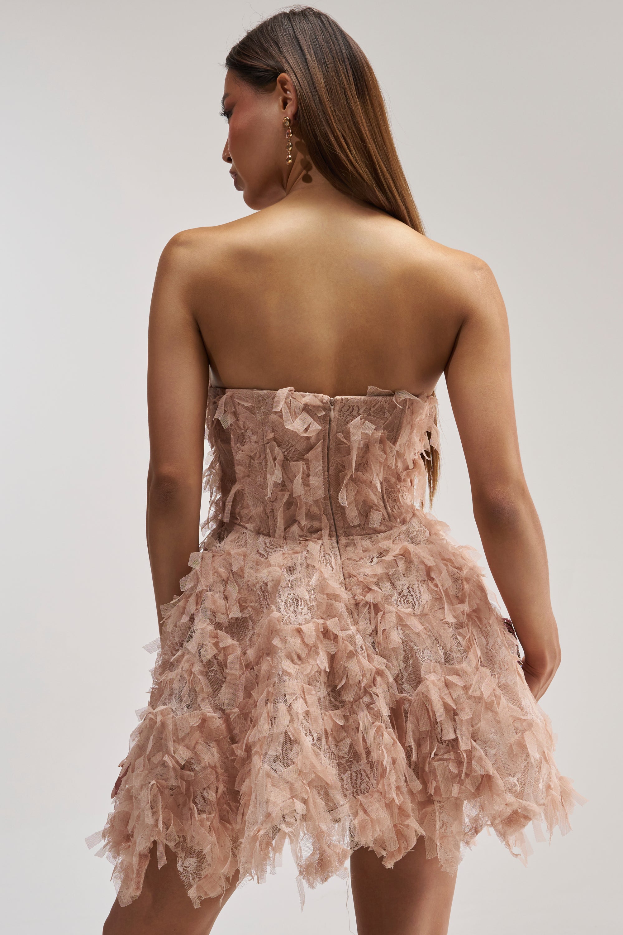 A woman with long brown hair faces away from the camera against a plain background, wearing the PRETTY PLEASE FLOCKED MINI DRESS IN MAUVE featuring a strapless, corset-style bodice and a ruffled skirt.