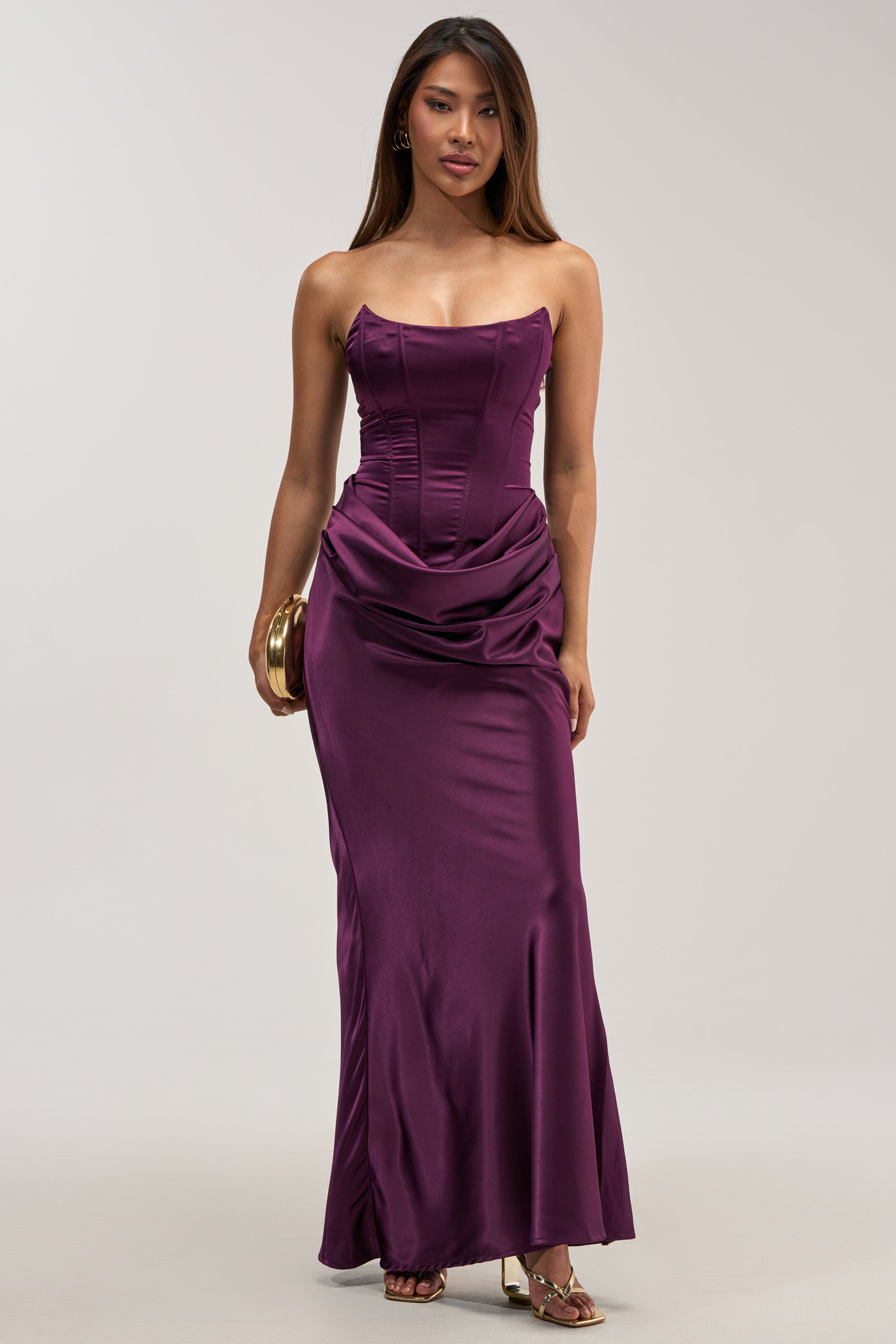 Clad in the HIGH STAKES SATIN MAXI DRESS—a strapless, rich purple satin gown featuring a structured bodice and elegantly draped skirt—a woman poses against a neutral backdrop, accessorizing with a gold clutch and gold sandals.