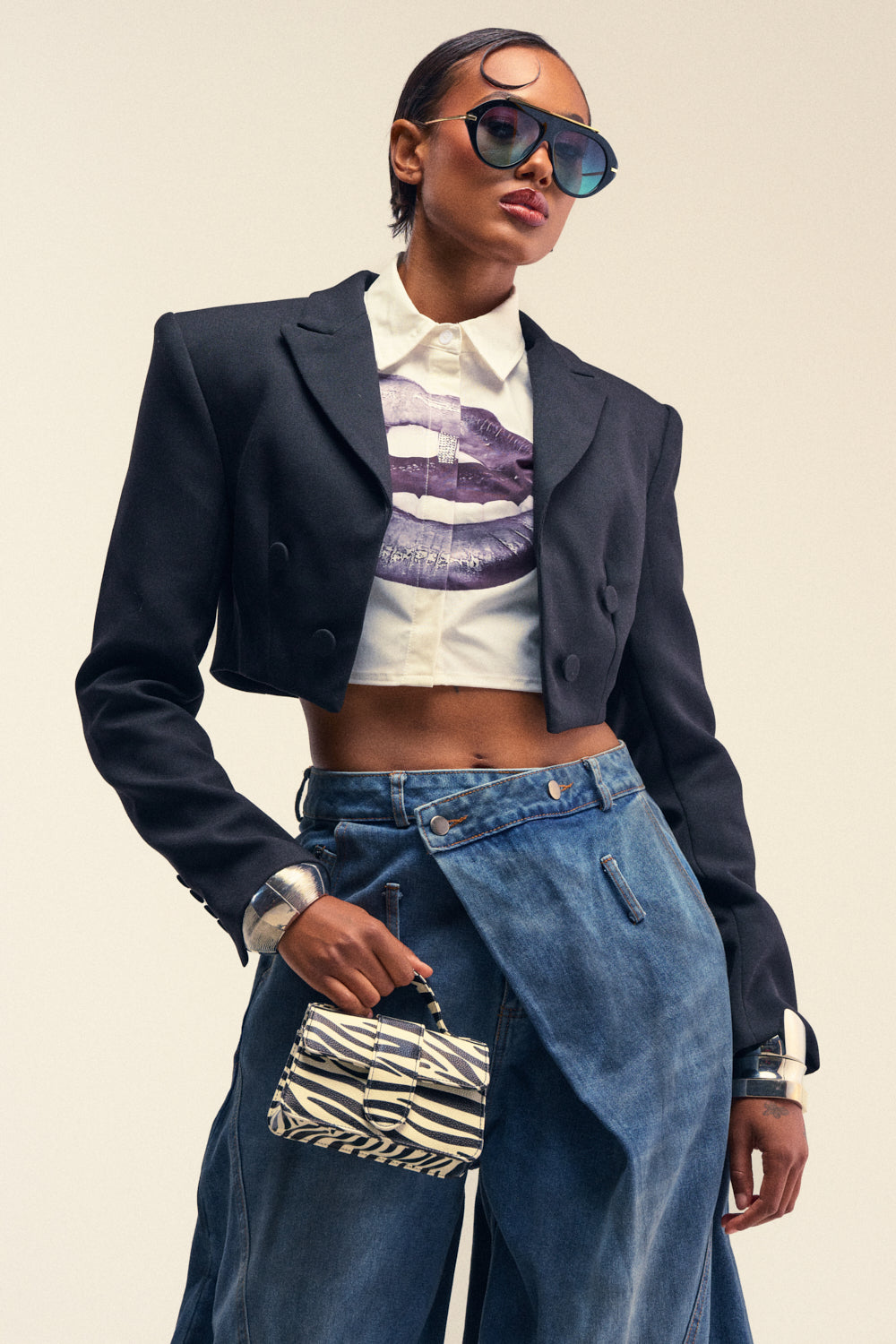Wearing the MAKE IT LOOK EASY STRUCTURED BLAZER, a stylish person poses confidently against a plain background. They complete the look with a graphic shirt, wide-leg jeans, sunglasses, and a striped mini handbag.