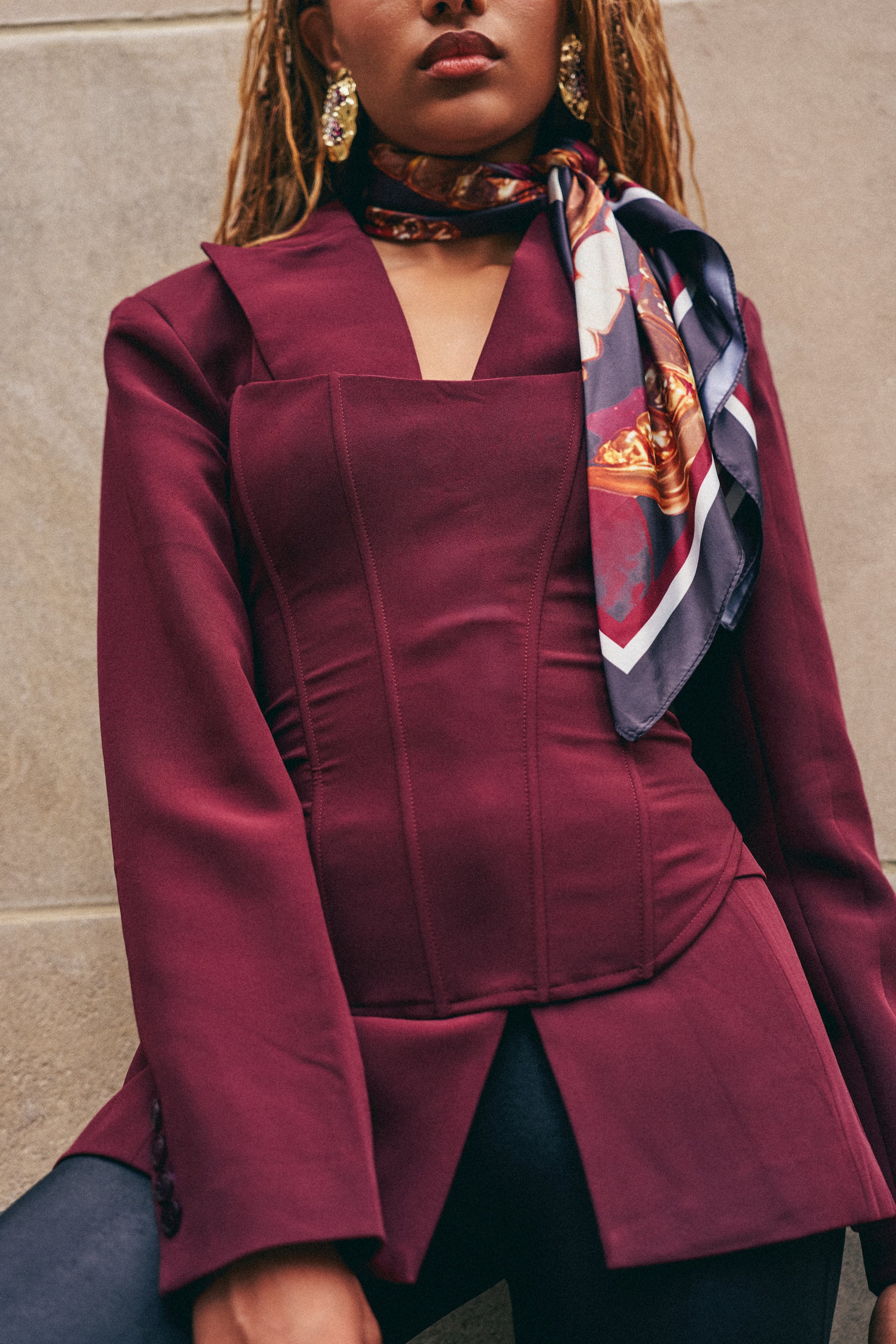 Wearing the EVERYDAY LUXE CORSET IN BURGUNDY beneath a chic maroon blazer, paired with a patterned silk scarf, black pants, and gold earrings, a person stands against a stone wall. Their face is partially out of frame.