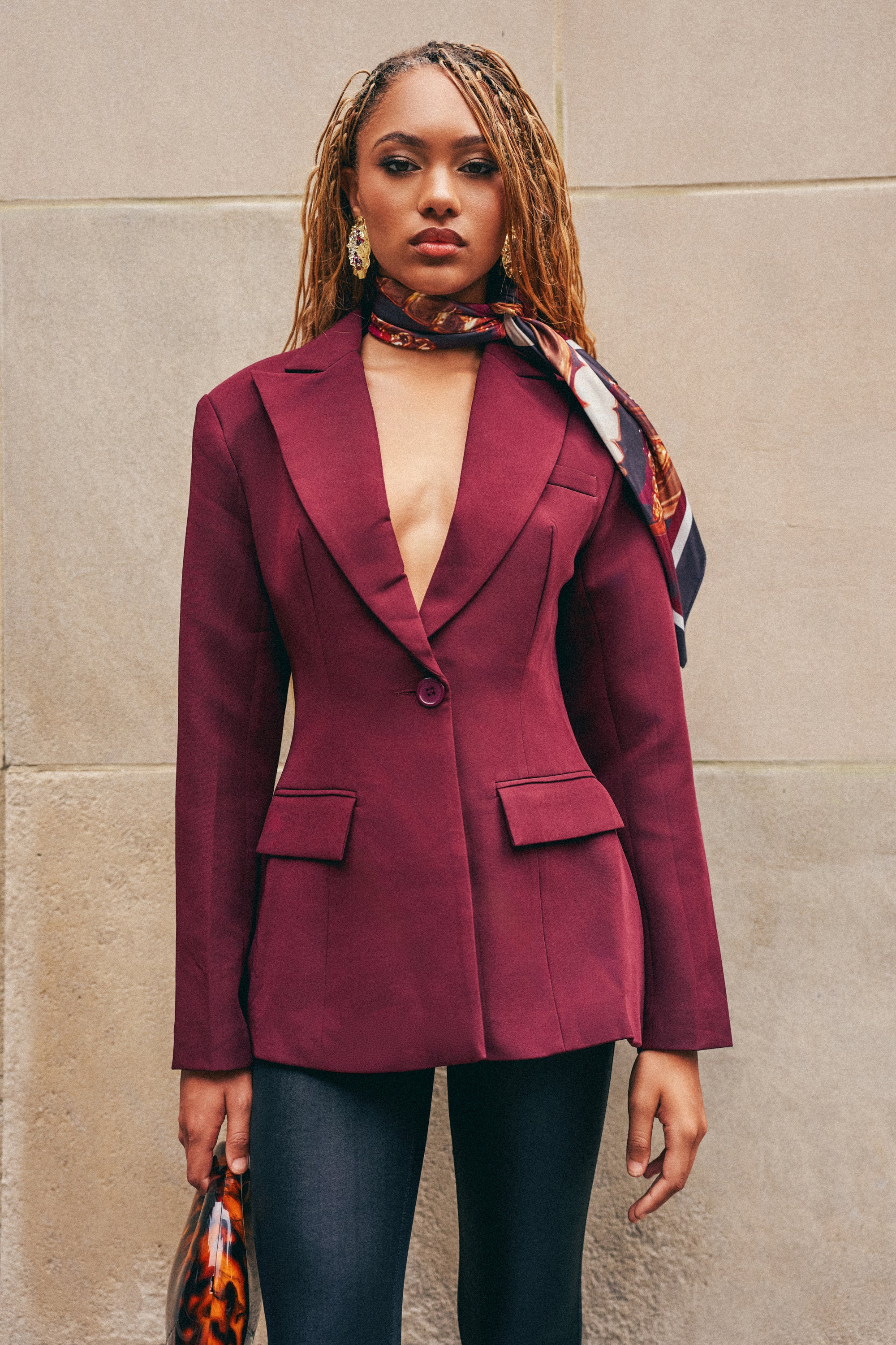 Wearing the EVERYDAY LUXE BLAZER IN BURGUNDY, a woman with long braids pairs it with dark pants and a patterned scarf draped around her neck, creating a sophisticated power dressing look. Against a beige stone wall and holding a small bag, she exudes modern workwear elegance.