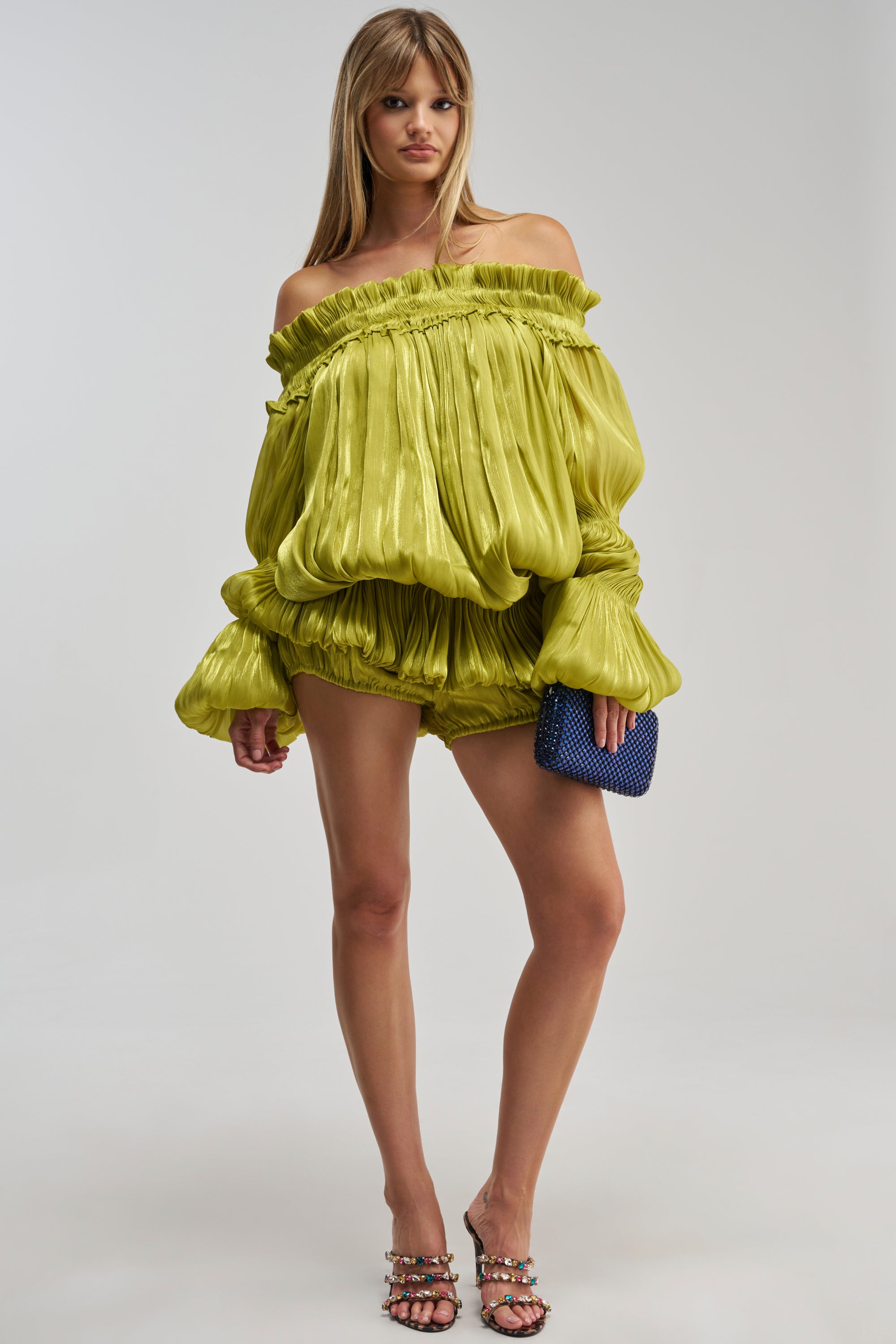 Wearing the BEAUTY QUEEN HOT SHORT in chartreuse, paired with an off-the-shoulder, lime green billowy blouse, this woman showcases one of the season’s standout statement looks. Her party-ready styling is finished with a small blue clutch and strappy heels as she stands confidently against a plain background.