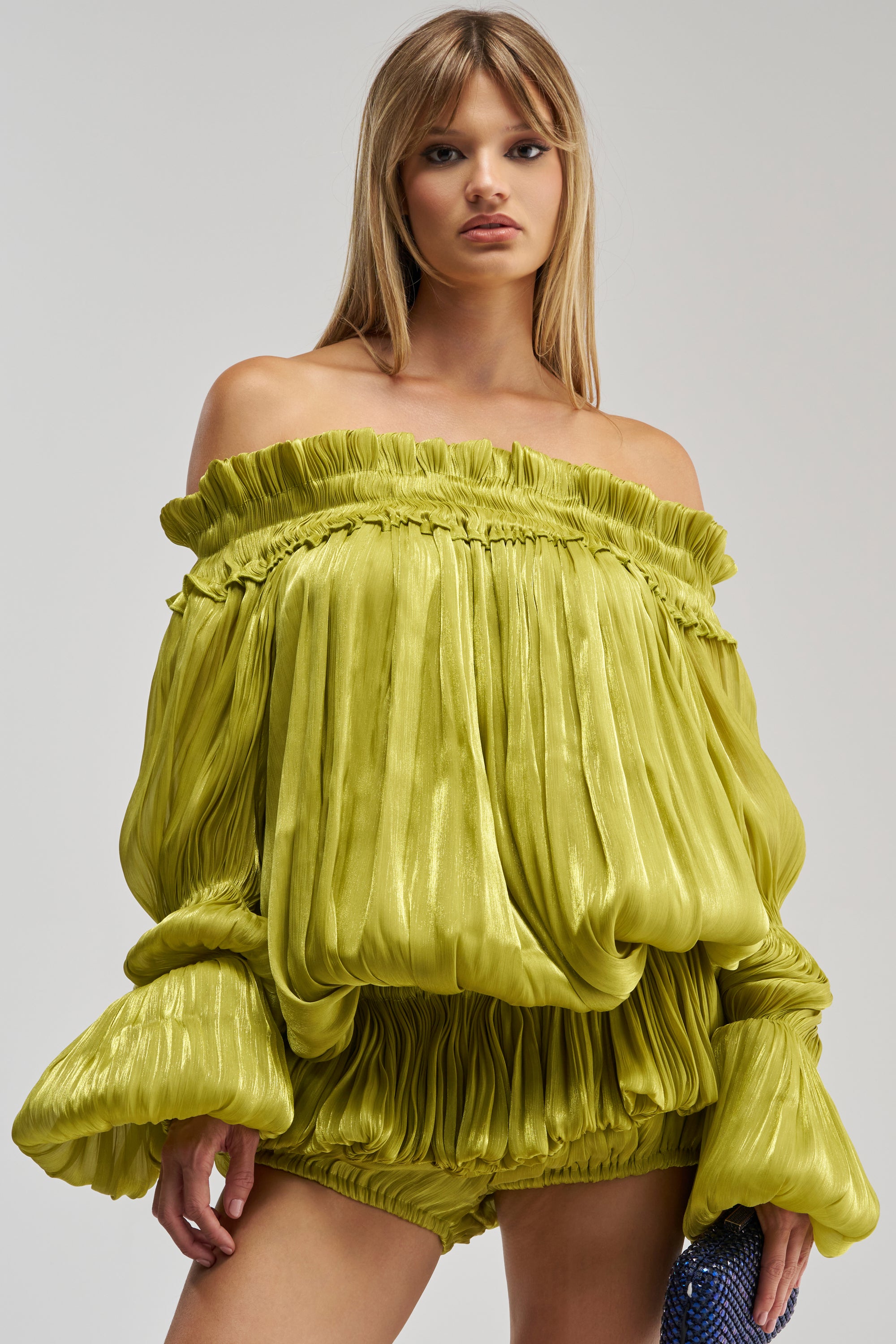 A woman wearing the BEAUTY QUEEN BLOUSE—with its shiny, off-the-shoulder satin-effect design, voluminous sleeves, and ruffled peplum hem—paired with matching lime green pleated shorts, stands against a plain background.
