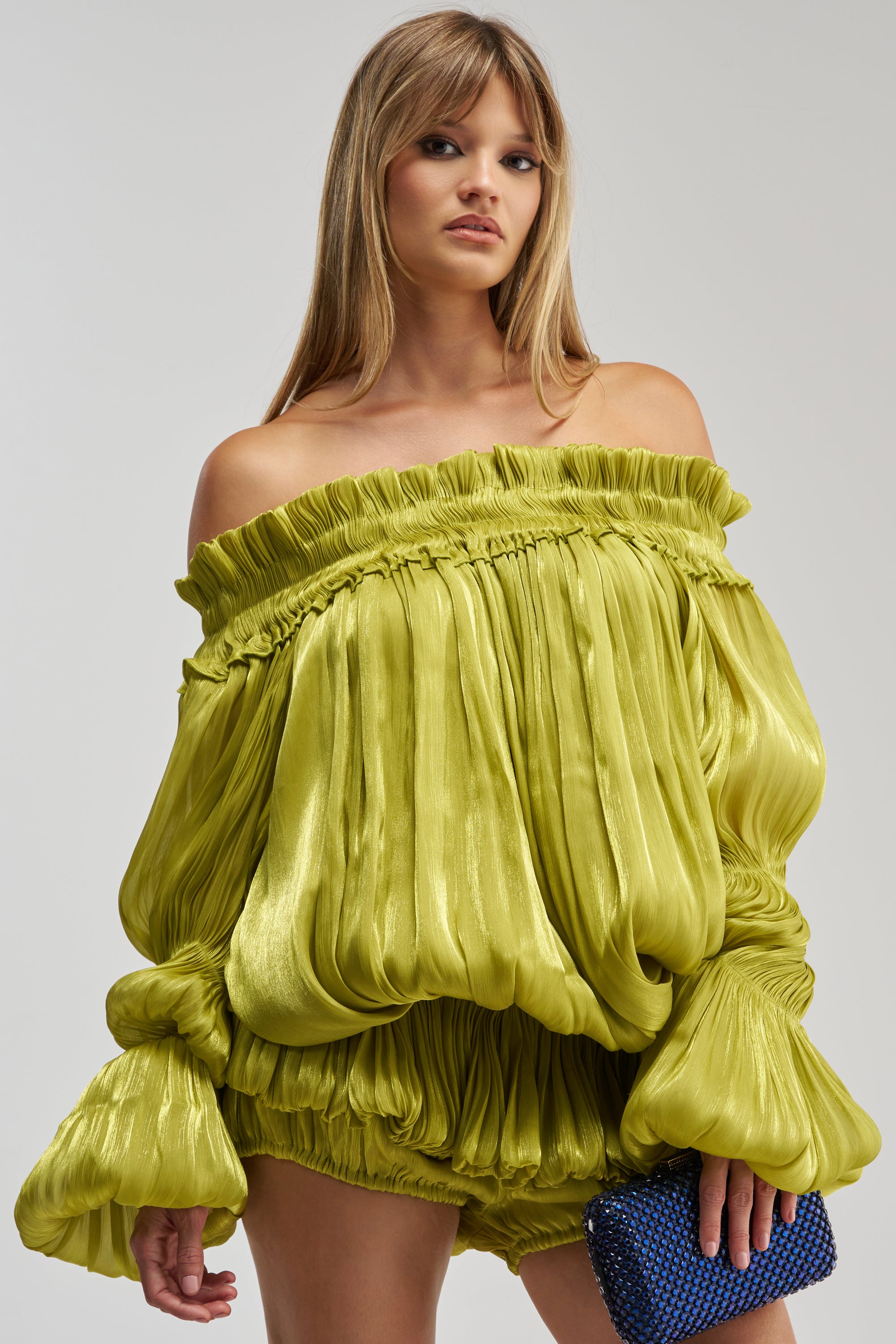 A woman wearing the BEAUTY QUEEN BLOUSE—a shiny, satin-effect, off-the-shoulder, pleated lime green top—paired with matching shorts and holding a small blue textured clutch.