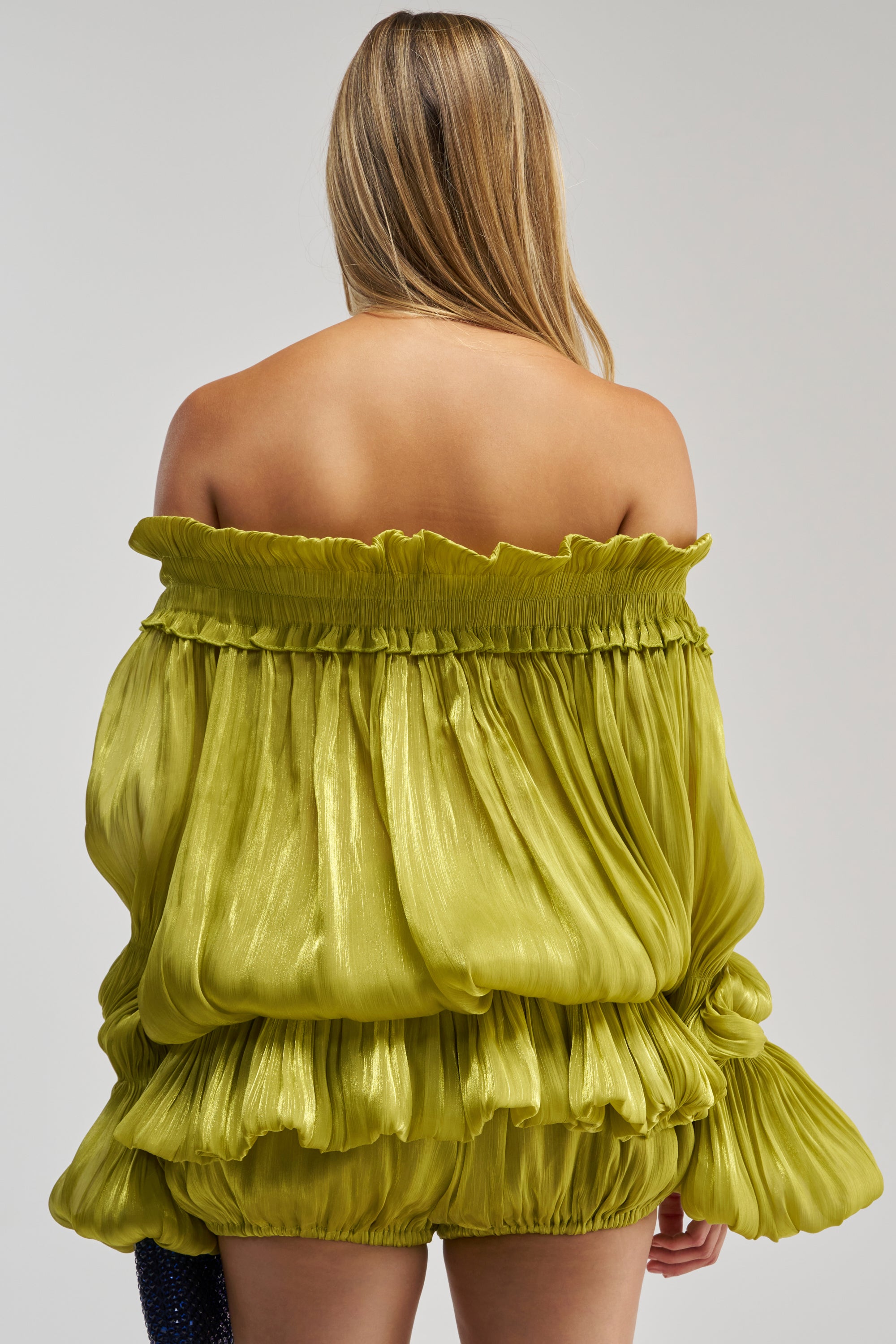 A person with long, blonde hair is seen from the back against a plain light background, wearing the BEAUTY QUEEN BLOUSE—an off-shoulder, shiny lime green top featuring a ruffled peplum hem.