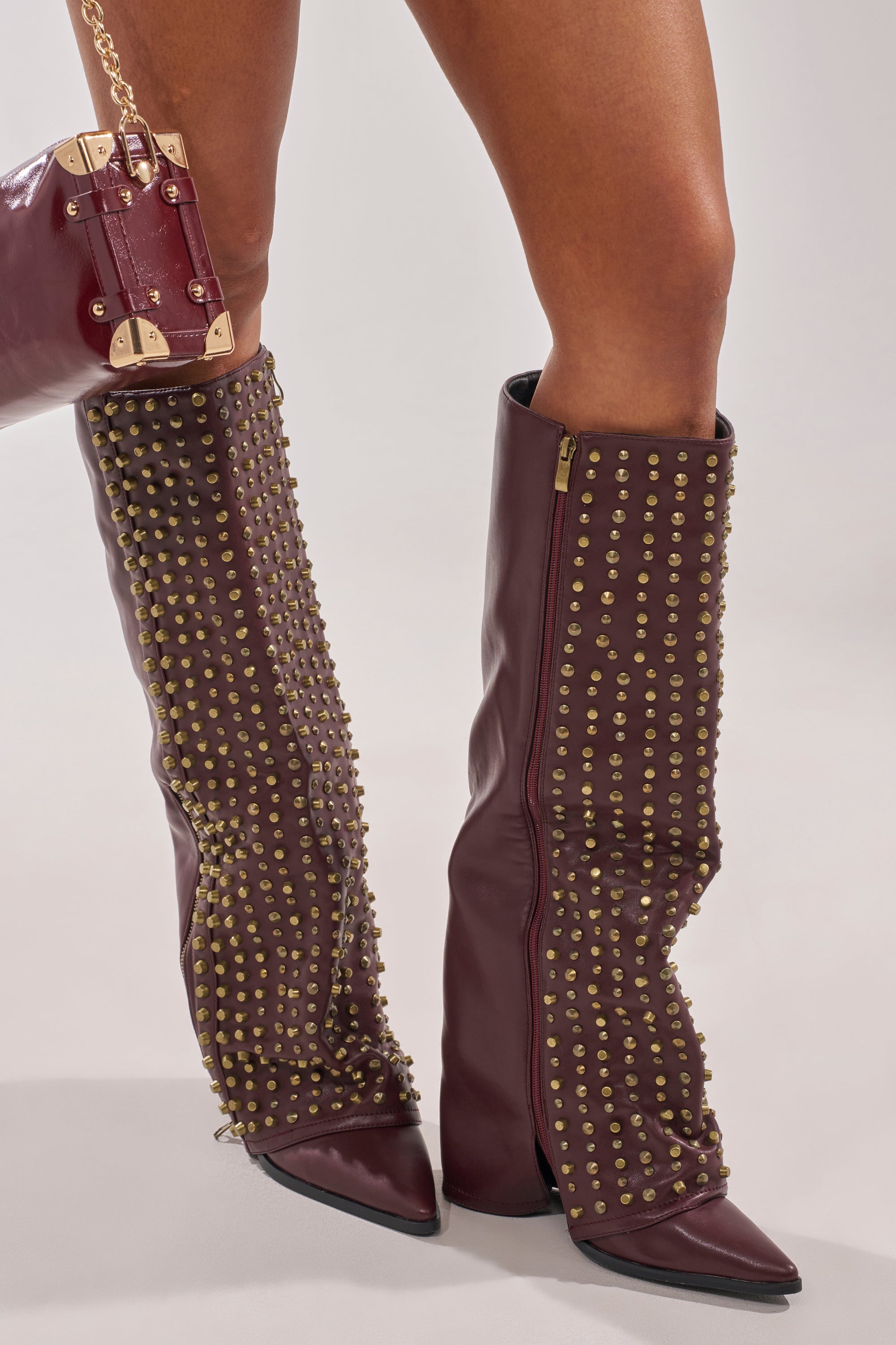 Wearing AZALEA WANG AGUSTUE Burgundy Studded Western Boots and a matching burgundy handbag with gold hardware, a person stands on a light-colored surface. 0