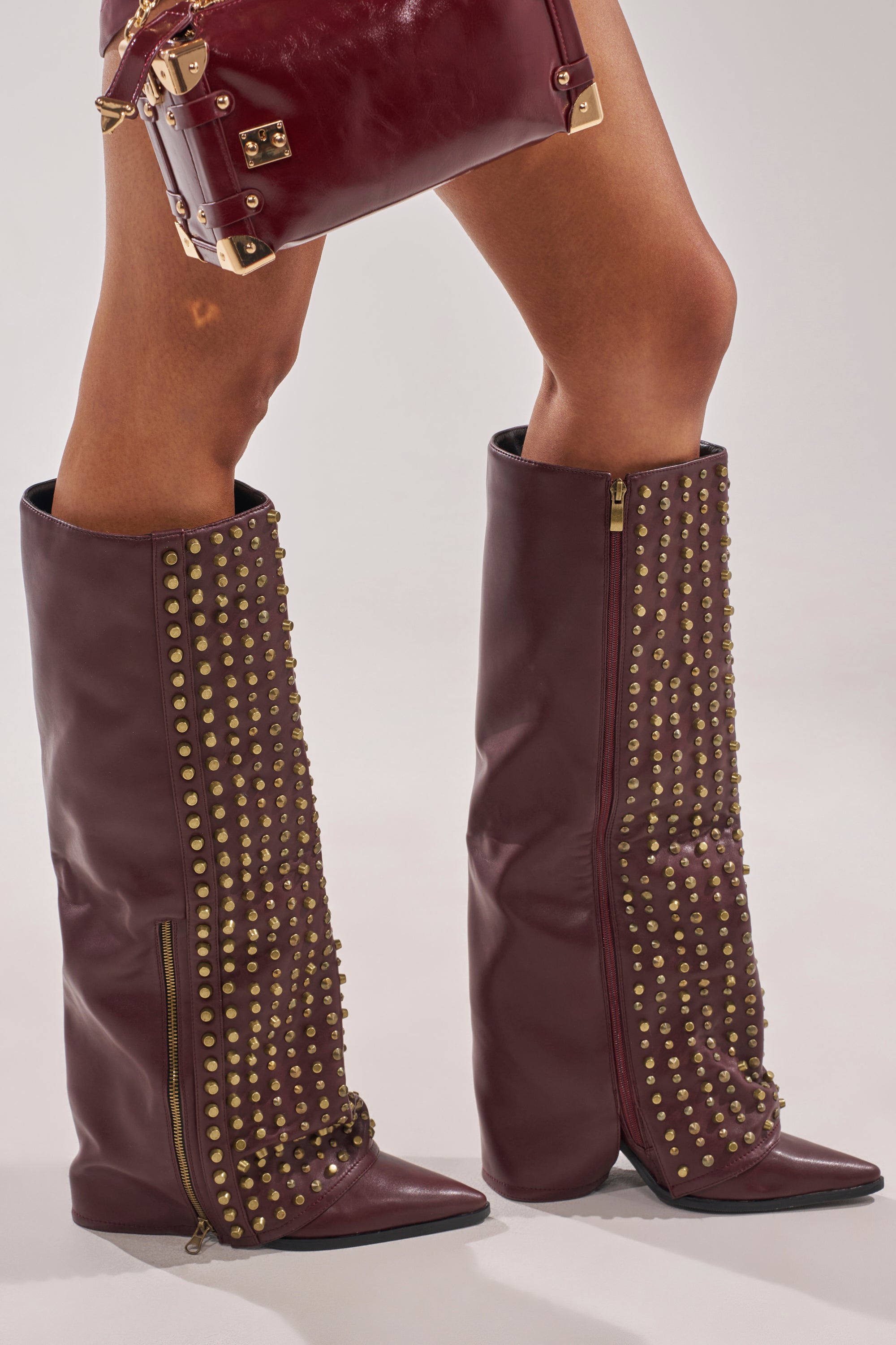 Wearing the AZALEA WANG AGUSTUE BURGUNDY STUDDED WESTERN BOOT, embellished with gold studs and zippers, and holding a coordinating burgundy handbag with gold hardware creates a bold, fashionable statement. 2