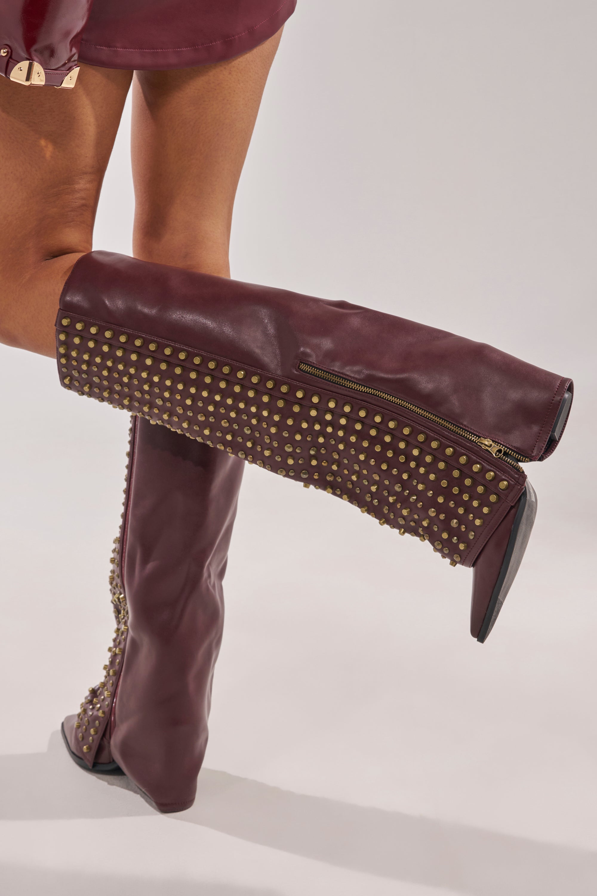 Someone crosses one leg over the other against a plain background, wearing the AZALEA WANG AGUSTUE BURGUNDY STUDDED WESTERN BOOT featuring gold studs and side zippers. 3