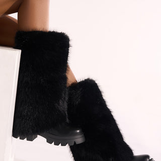 A person sits cross-legged, highlighting the AZALEA WANG AHSOKA BLACK FUR FLATFORM BOOT with chunky soles. The knee-high design and fuzzy texture stand out against a plain, light background.