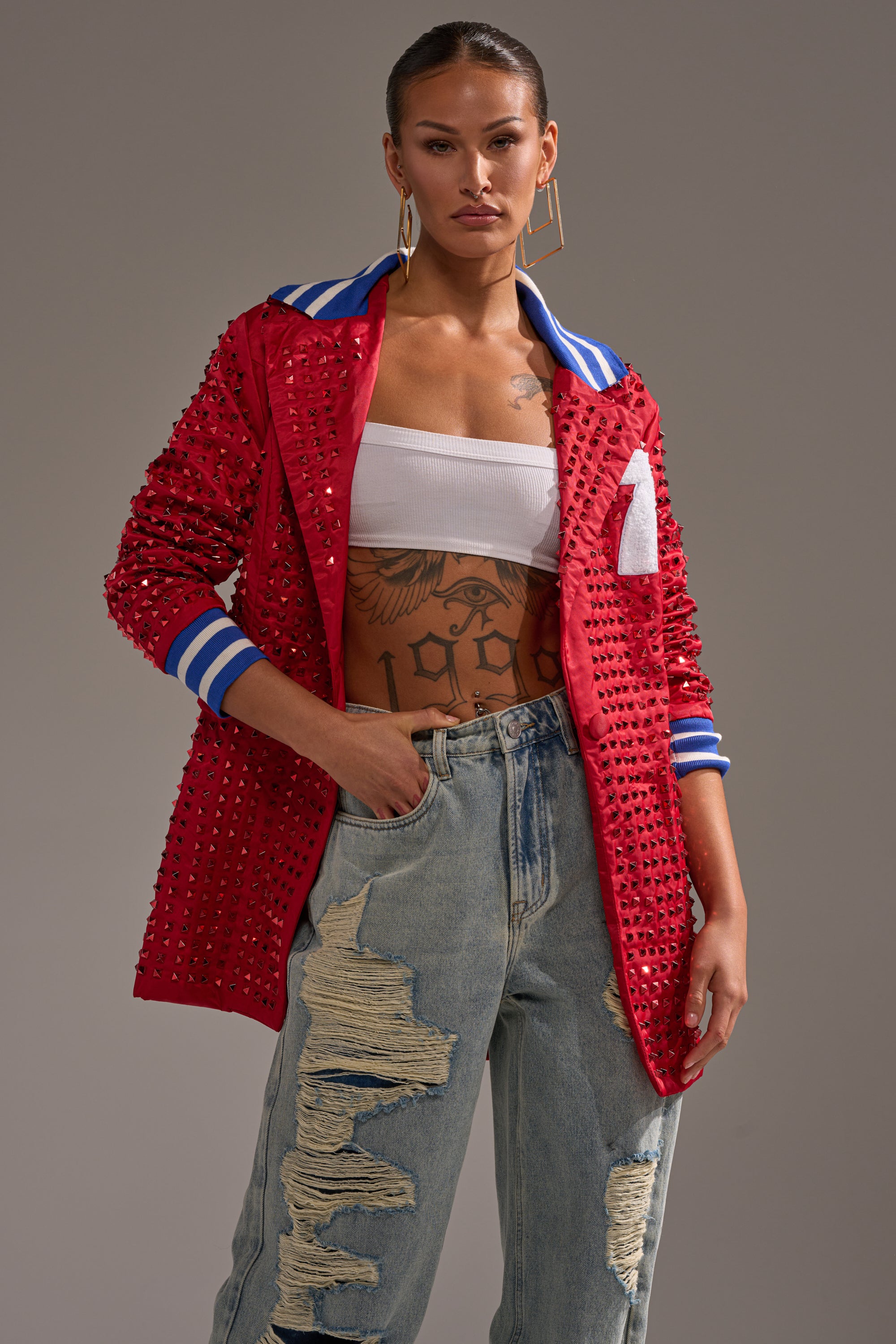 A woman stands confidently in the FIRST ROUND DRAFT PICK STUDDED BLAZER IN RED with blue and white striped cuffs, styled with a white bandeau top, ripped jeans, gold hoop earrings, and a large chest tattoo.